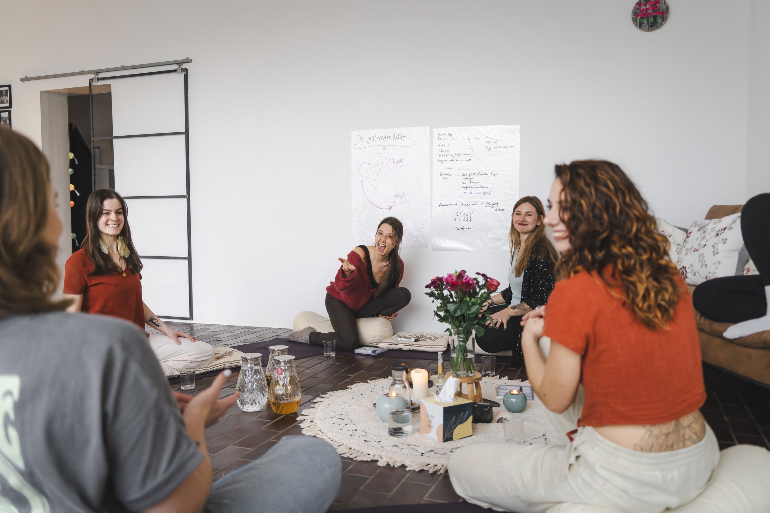 Eine Gruppe von Frauen sitzt in einem Kreis auf dem Boden in einem Frauenzirkel. Vor ihnen befinden sich Kerzen, Blumen und Trinkgläser auf einem Tischset. Im Hintergrund hängen Plakate an der Wand.