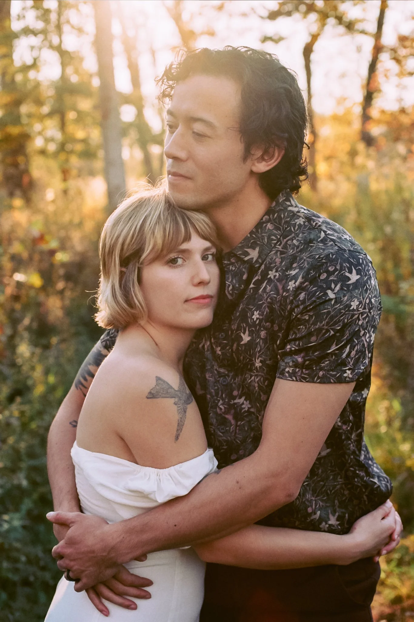 A couple hugging outdoors during sunset, surrounded by autumn trees.