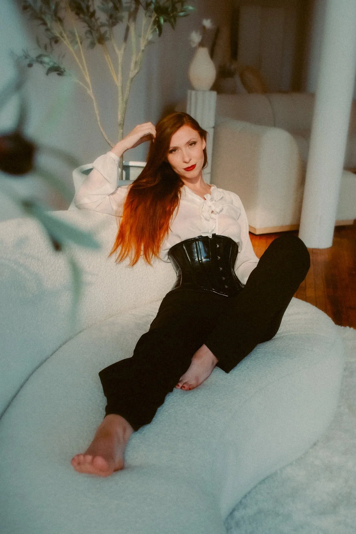 A woman with long red hair sitting on a white sofa, wearing a white blouse, black pants, and a shiny black corset, in a cozy living room setting.