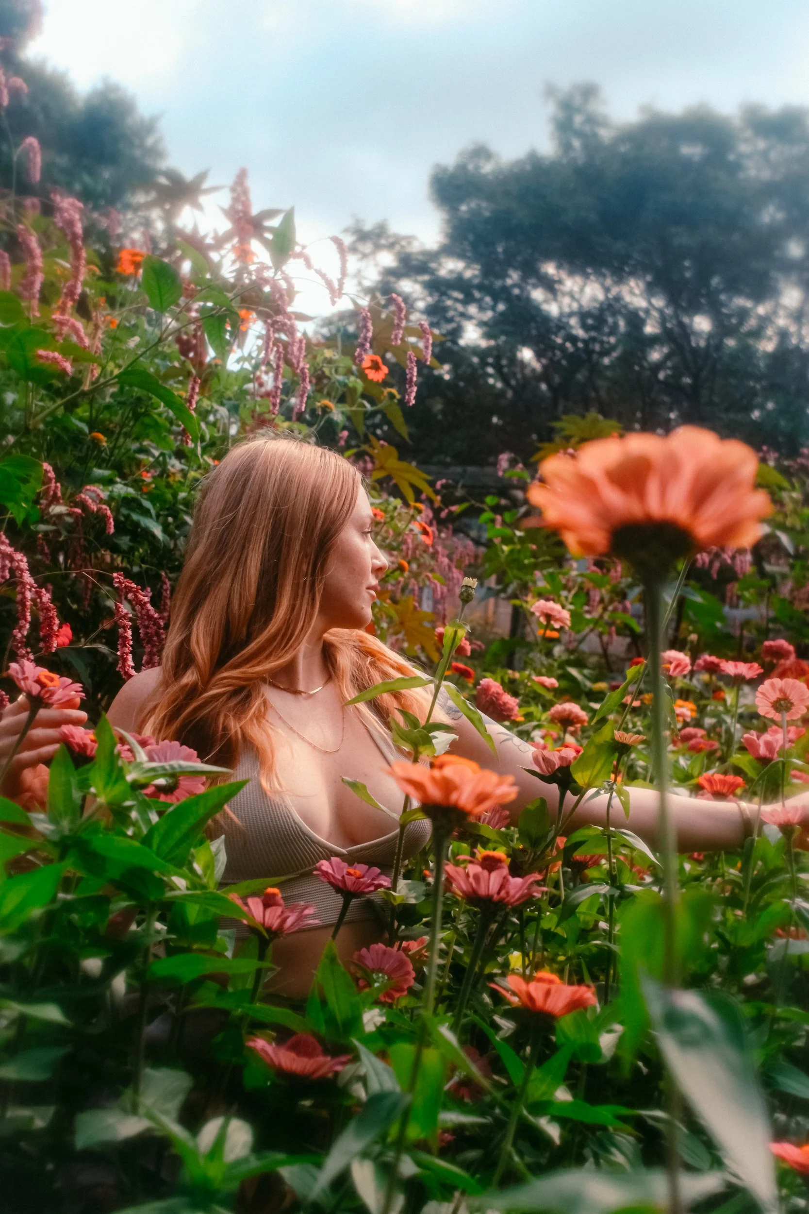 A woman with long red hair among pink and orange flowers in a garden with trees and sky in the background.