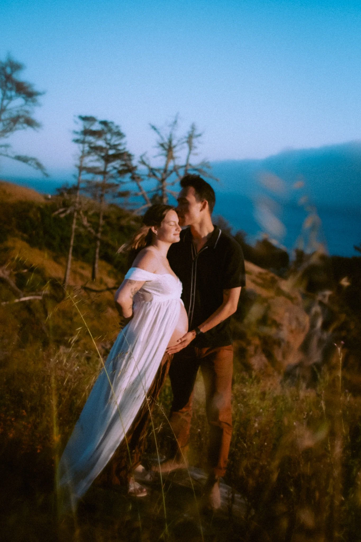 A pregnant woman and a man standing outdoors at sunset, holding hands over her belly, with a scenic landscape of trees and mountains in the background.