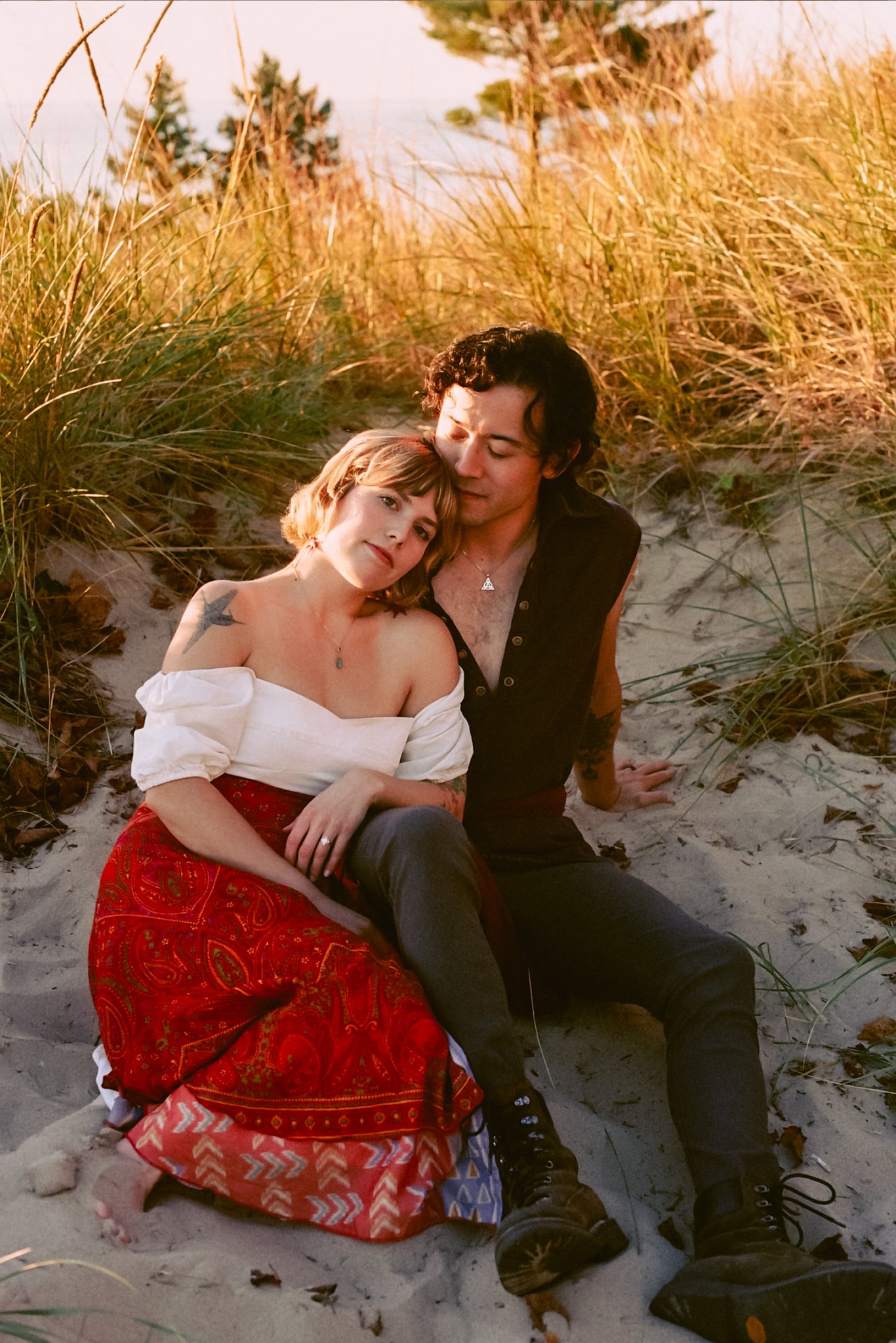 A young couple sitting closely together on the sand among tall grass at sunset, with the woman resting her head on the man's shoulder.
