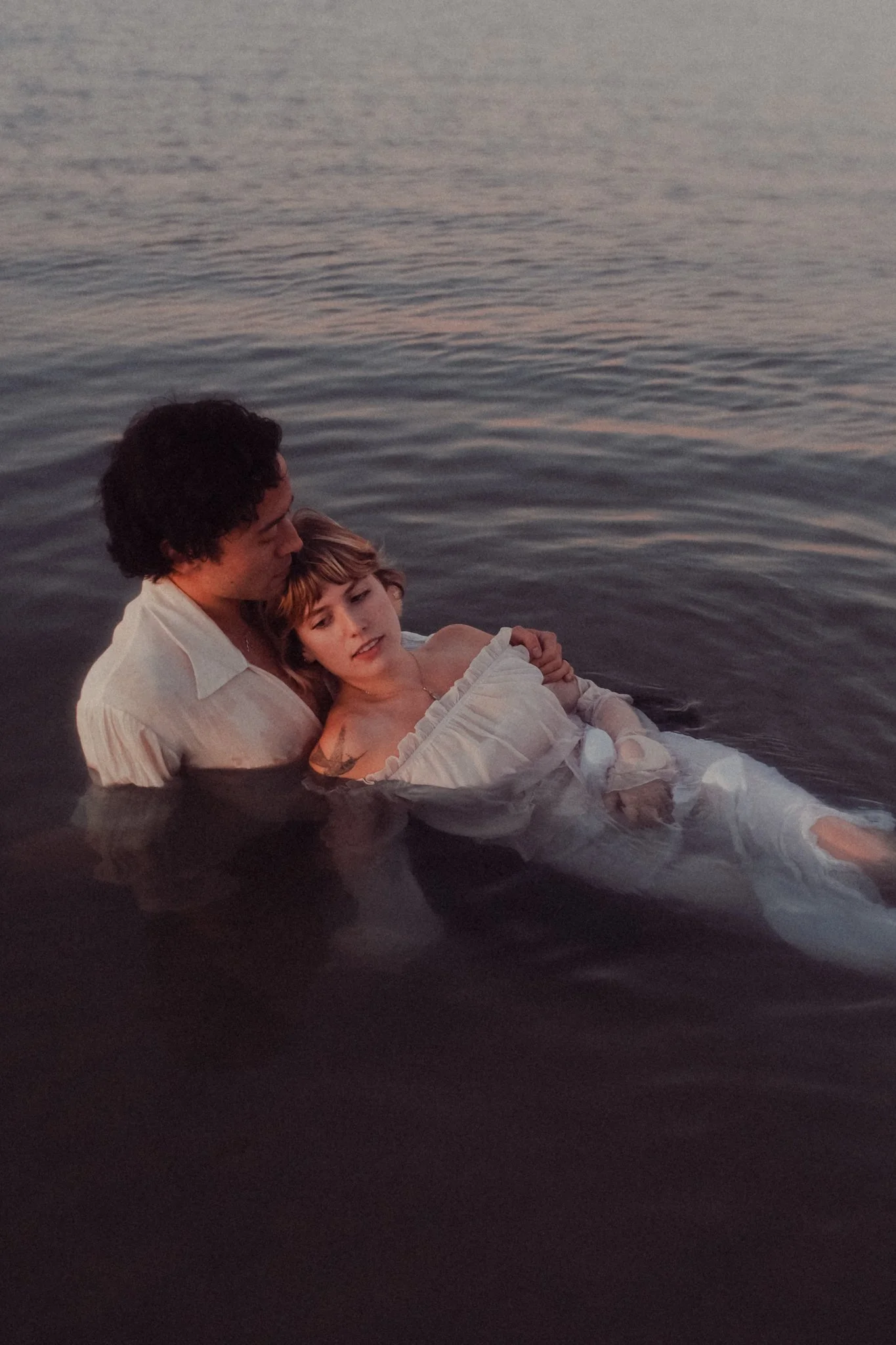 A person holding a woman in white clothing in calm water during sunset or dusk.