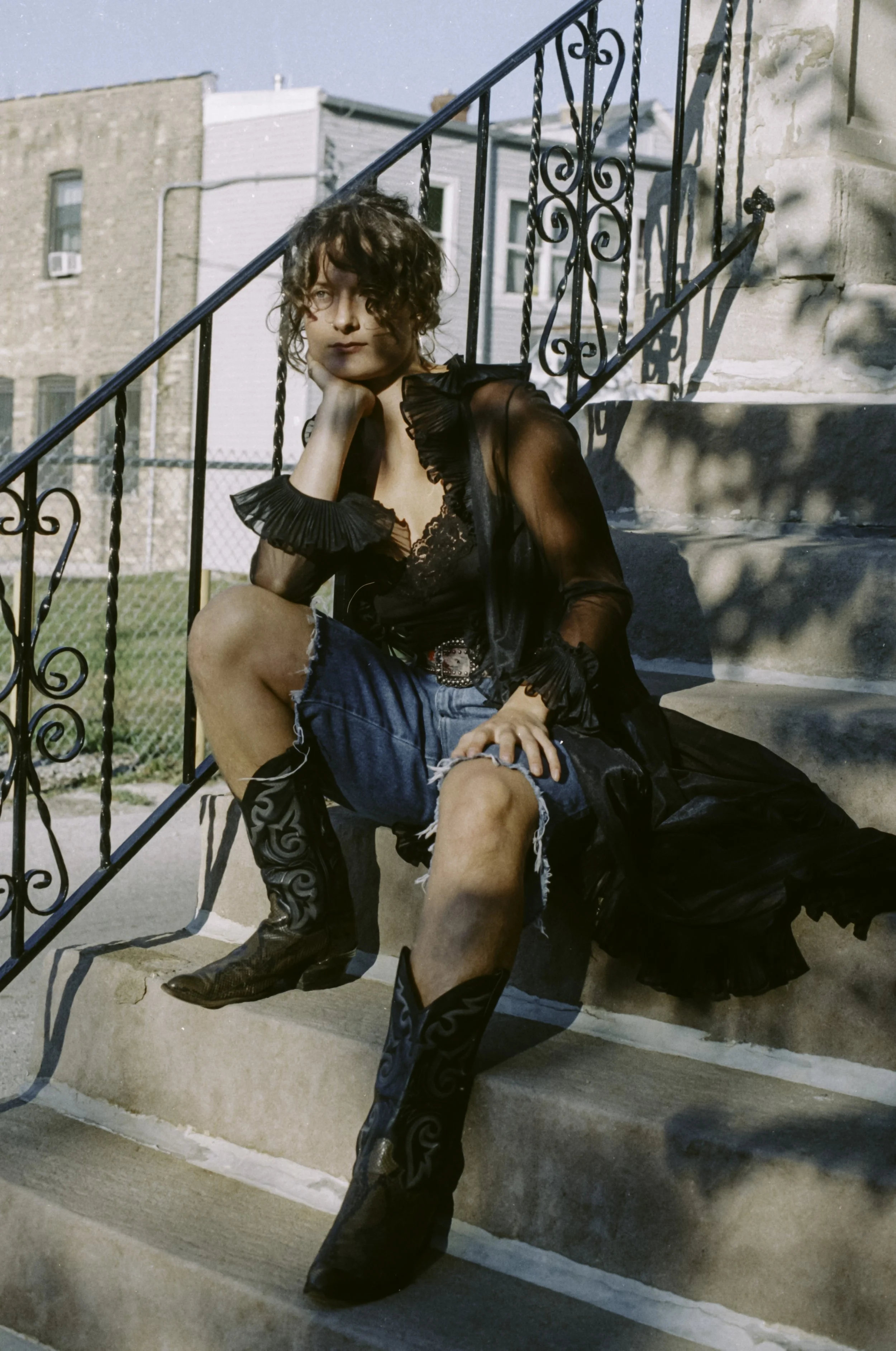 A young person with curly hair sitting on outdoor concrete stairs, wearing a sheer black blouse, tuxedo boots, denim shorts, and a belt, resting their chin on their hand.