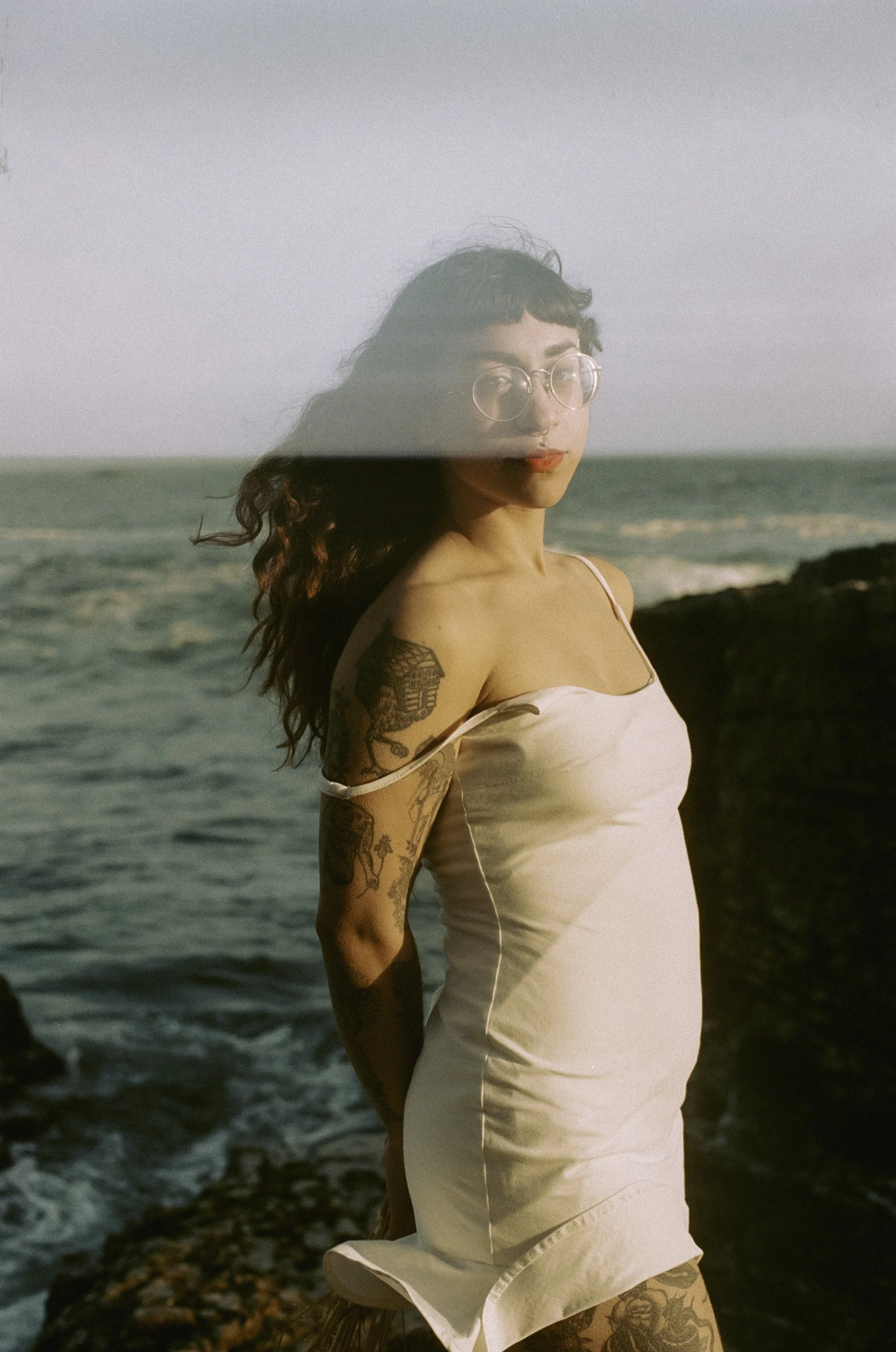 A woman standing on rocks near the ocean, wearing a white dress and glasses, with tattoos on her arm and leg, during sunset.