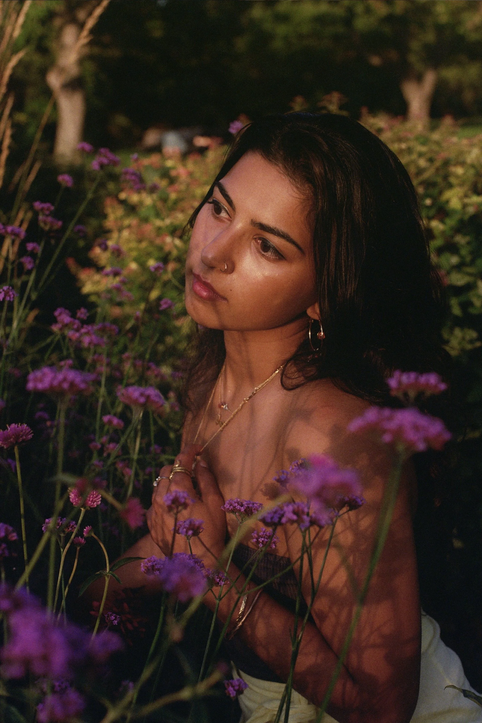 A young woman with dark hair and jewelry is sitting among purple flowers in a garden at sunset, looking contemplative.