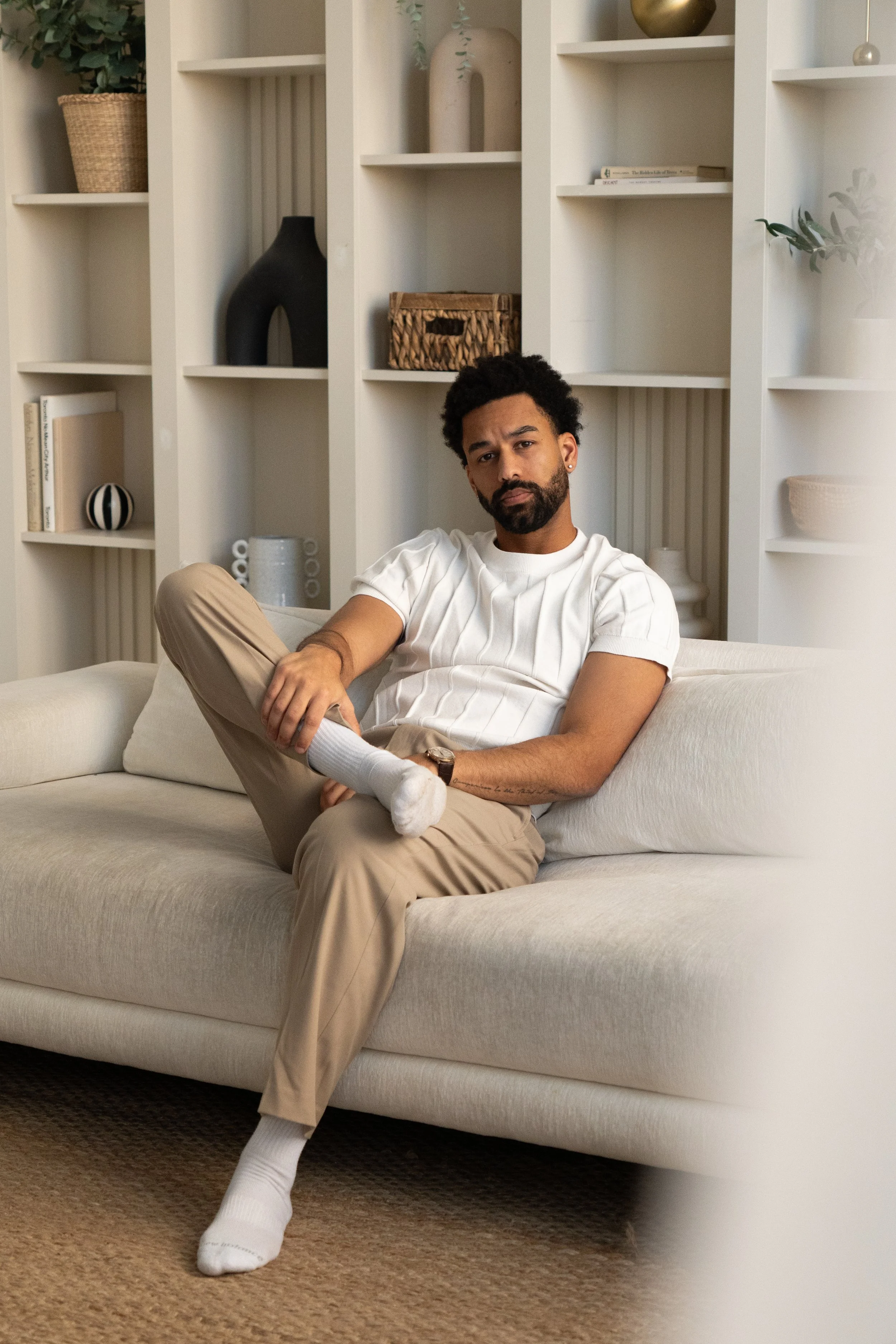A man sitting on a beige sofa with one leg crossed, wearing a white shirt, khaki pants, and white socks. Behind him is a white bookshelf with decorative items and books.