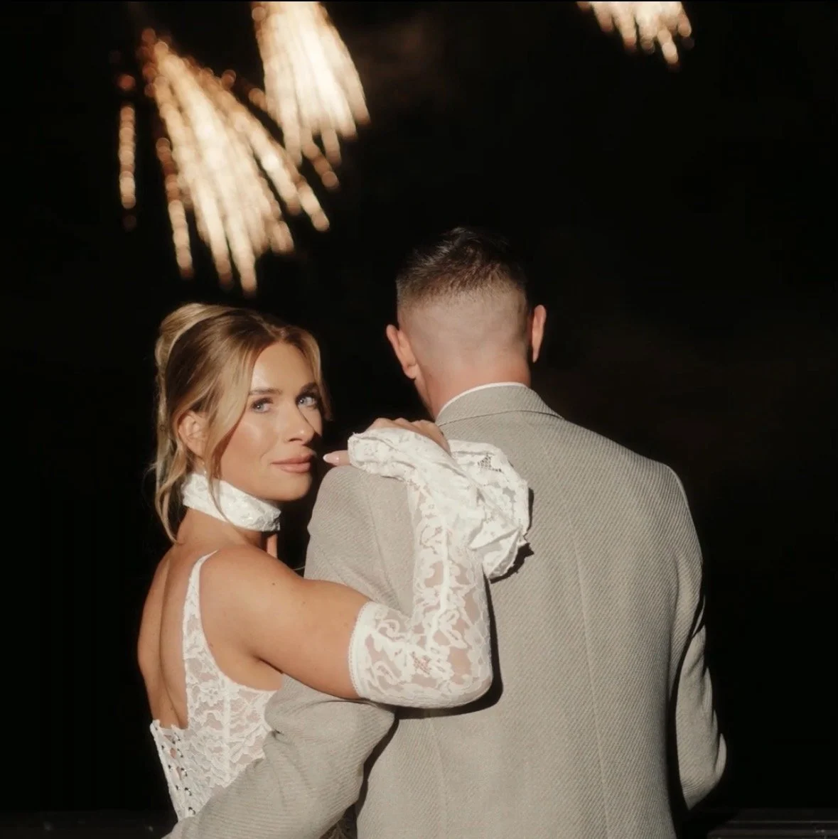 A woman in a wedding dress and gloves looks over her shoulder at a man in a gray suit, with fireworks in the background.