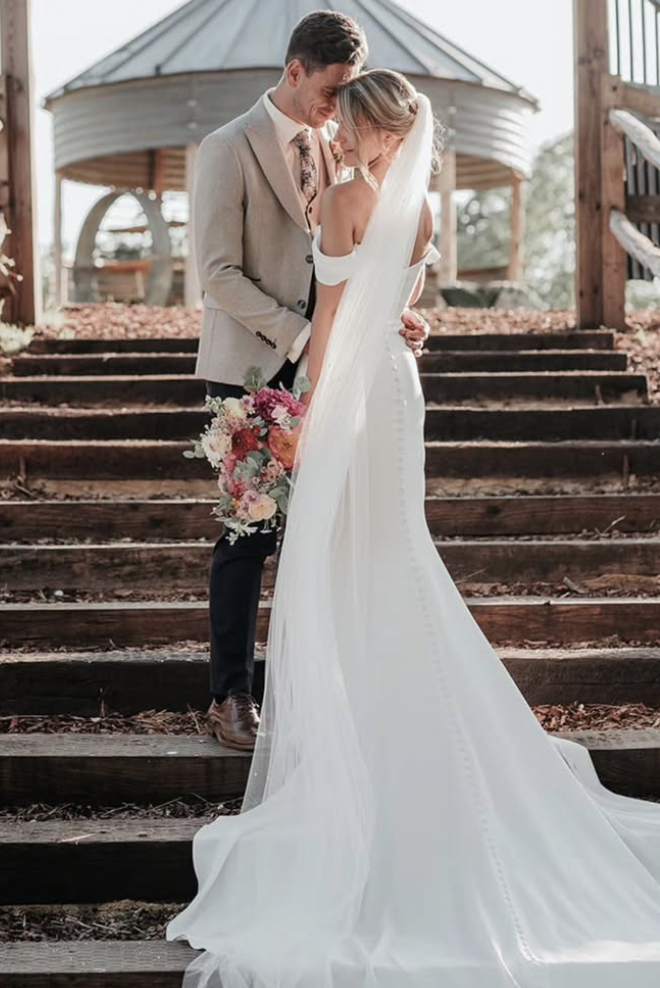 A bride and groom embrace on outdoor wooden stairs, with the bride wearing a white wedding gown and veil, holding a colorful bouquet, and the groom in a light gray suit with a floral tie, in a scenic rustic setting.