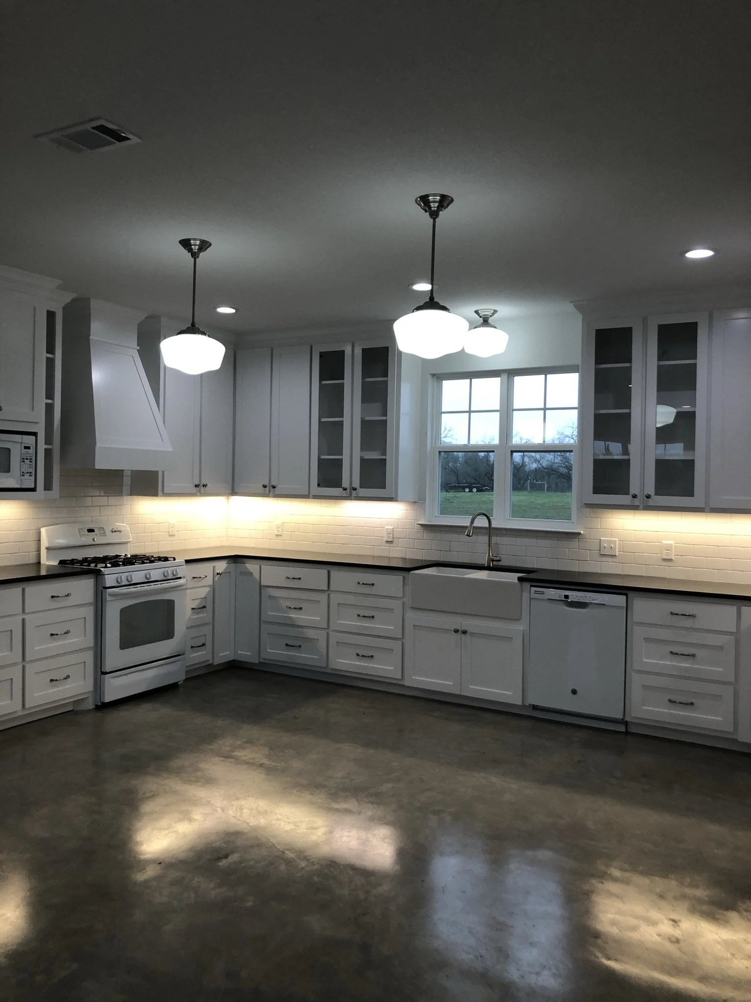 Modern kitchen with white cabinets, black countertops, white subway tile backsplash, and stainless steel appliances. Two pendant lights hang over the workspace, and a window above the sink offers a view of outside. The floor is polished concrete.
