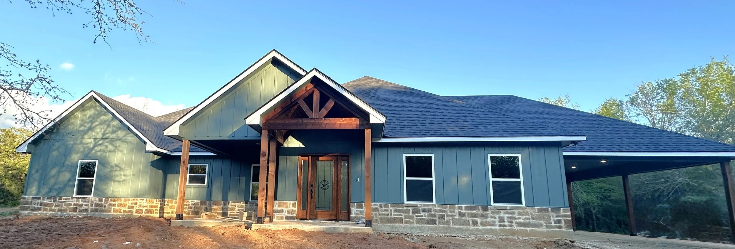 Newly constructed two-story house with blue exterior siding, stone foundation, wooden front porch with a gable roof, and surrounded by trees under a clear blue sky.