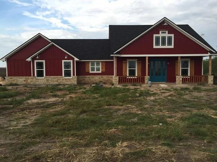 A newly built house with a red exterior, stone foundation, and a centered front porch with a blue door and railings, set against a partly cloudy sky.