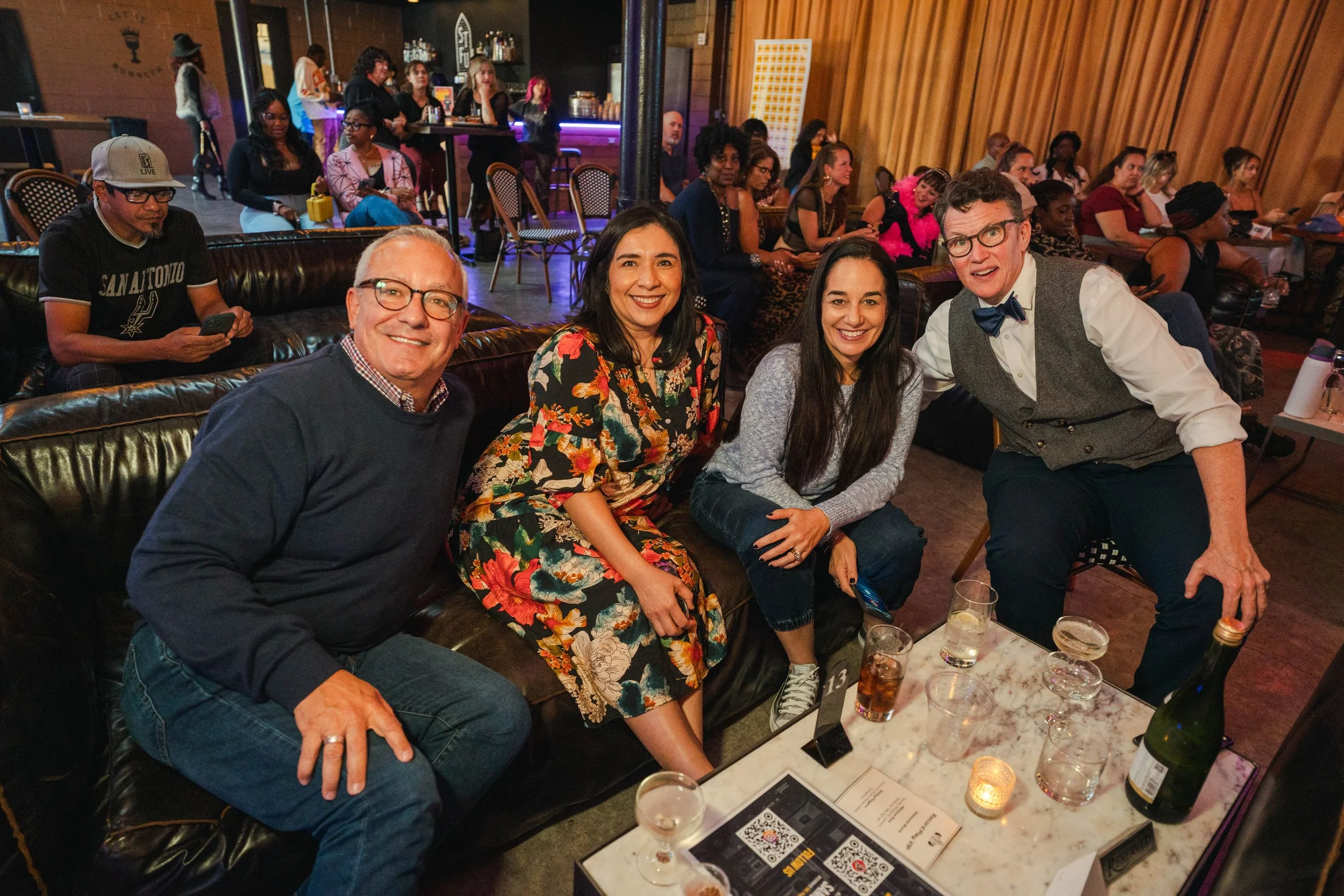 A group of four people sitting on a black leather couch at a social gathering. They are smiling at the camera, with drinks and a champagne bottle on the table in front of them. The background shows more people seated and standing in a dimly lit venue