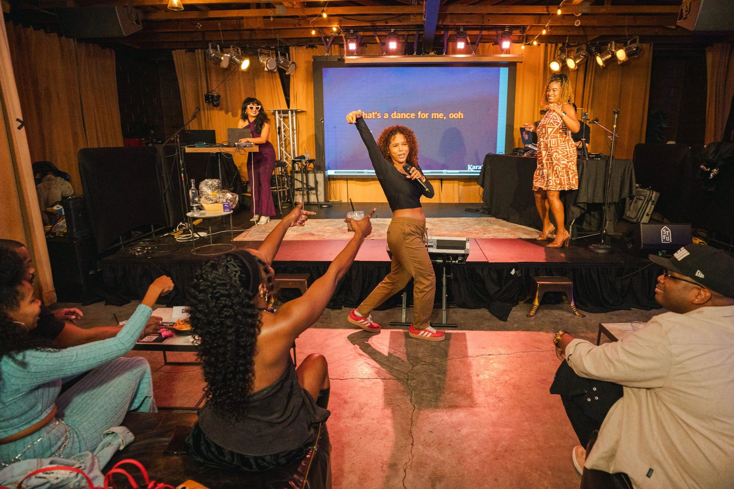 A woman singing and dancing with a microphone on stage at a karaoke event, with two women in the background using a DJ setup, and multiple audience members enjoying the performance in a cozy venue.