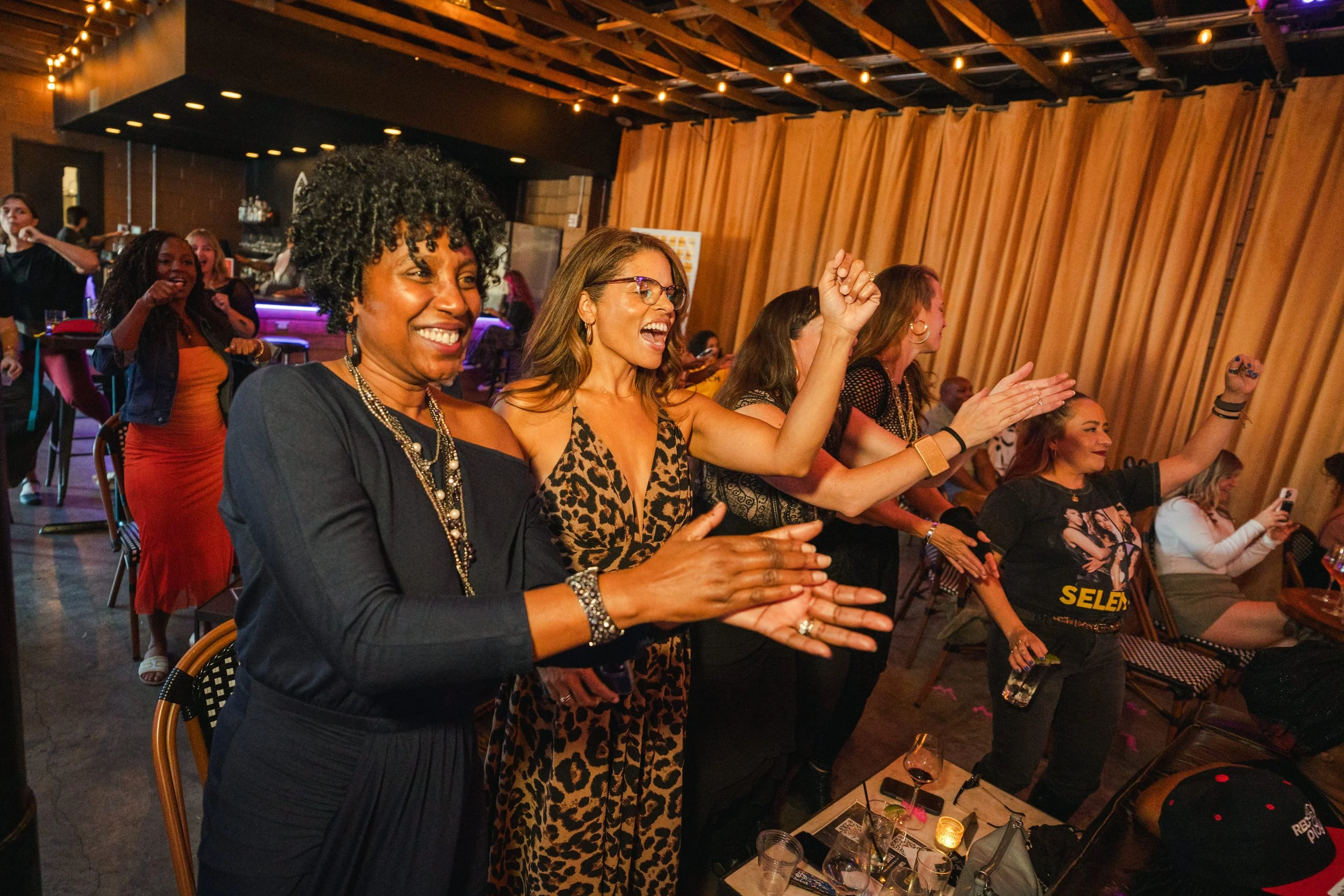 Group of women at a lively party, clapping, dancing, and smiling, with warm lighting and a wooden ceiling in the background.