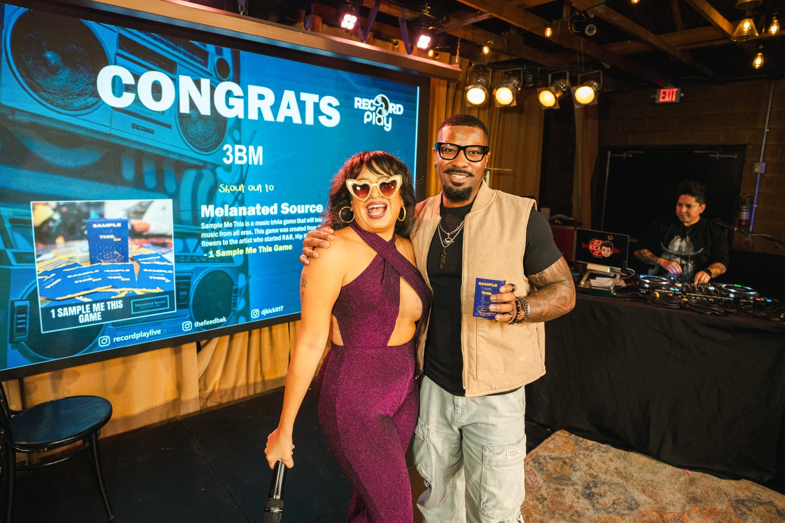 Two people standing together in a celebration setting, smiling at the camera, with a DJ in the background. The woman on the left wears heart-shaped sunglasses and a purple dress with cutouts. The man on the right wears glasses, a beige vest, and hold