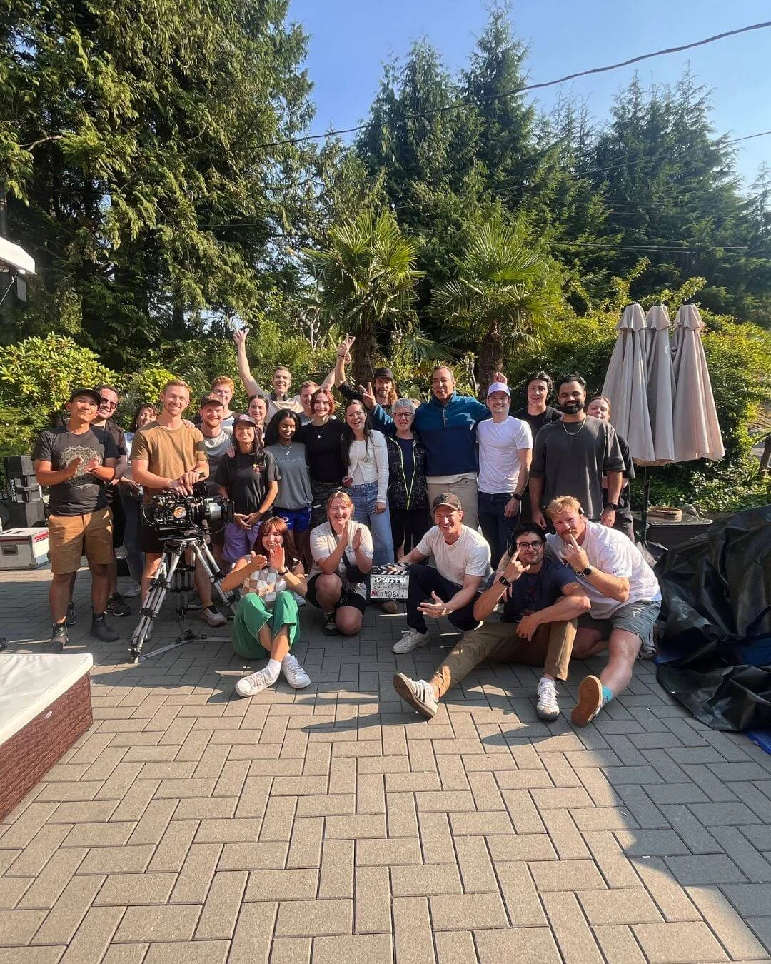 A large group of people gathered outdoors on a paved area, smiling and posing for a photo with trees and greenery in the background, and some umbrellas on the right side.