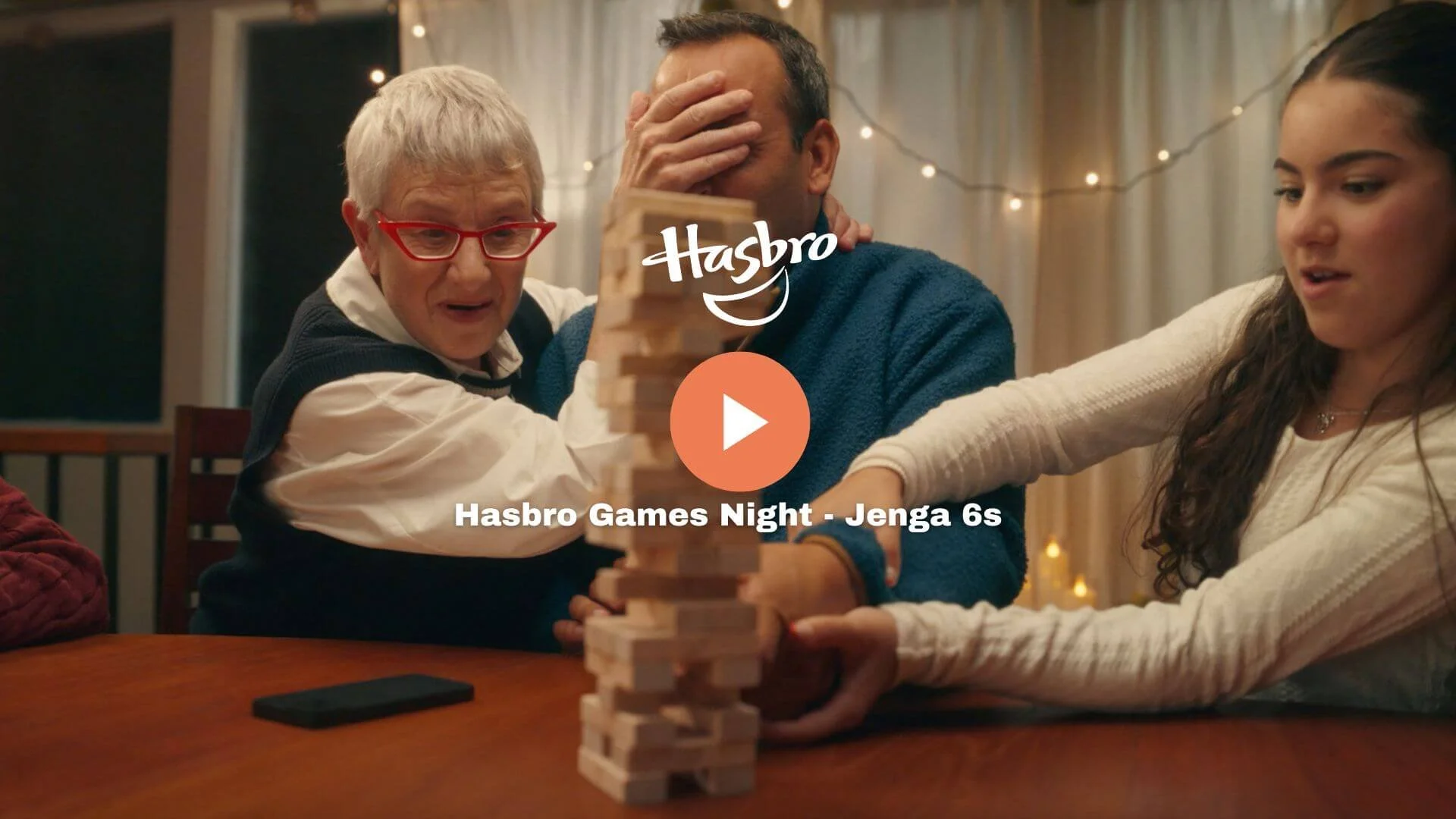 Three people playing Jenga, an elderly woman with red glasses, a man with a hand on his face, and a young woman, sitting around a table with a mobile phone, under warm lighting with string lights in the background.