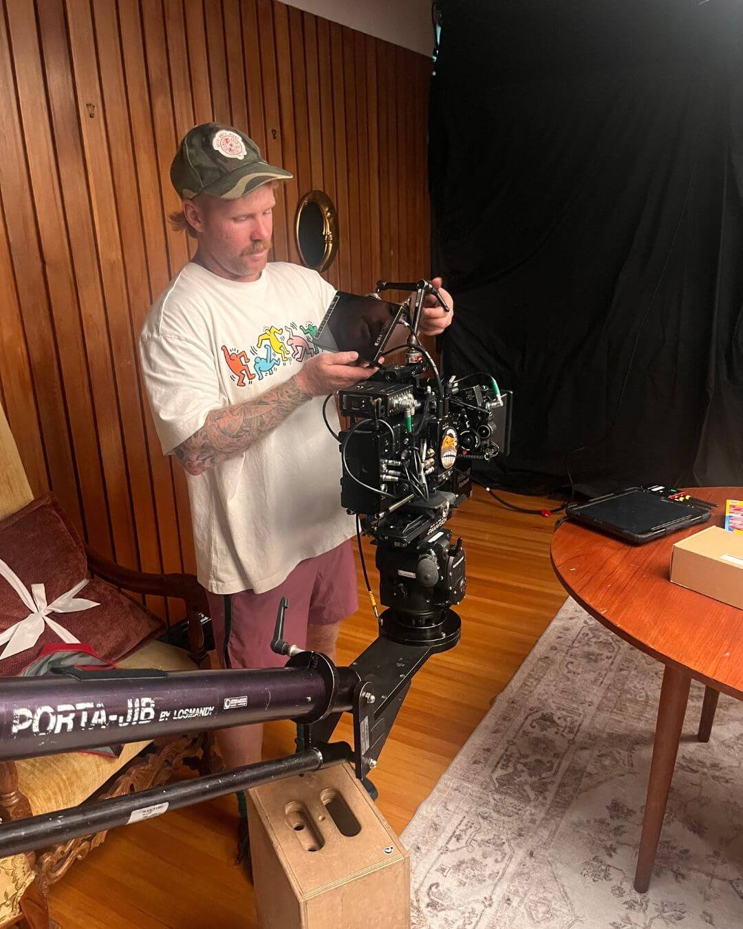 A man wearing a white t-shirt with colorful dancing figures and a camouflage baseball cap is operating a professional film camera on a studio set. The scene includes a wooden-paneled wall, a vintage wooden chair with a white ribbon, and a table with 