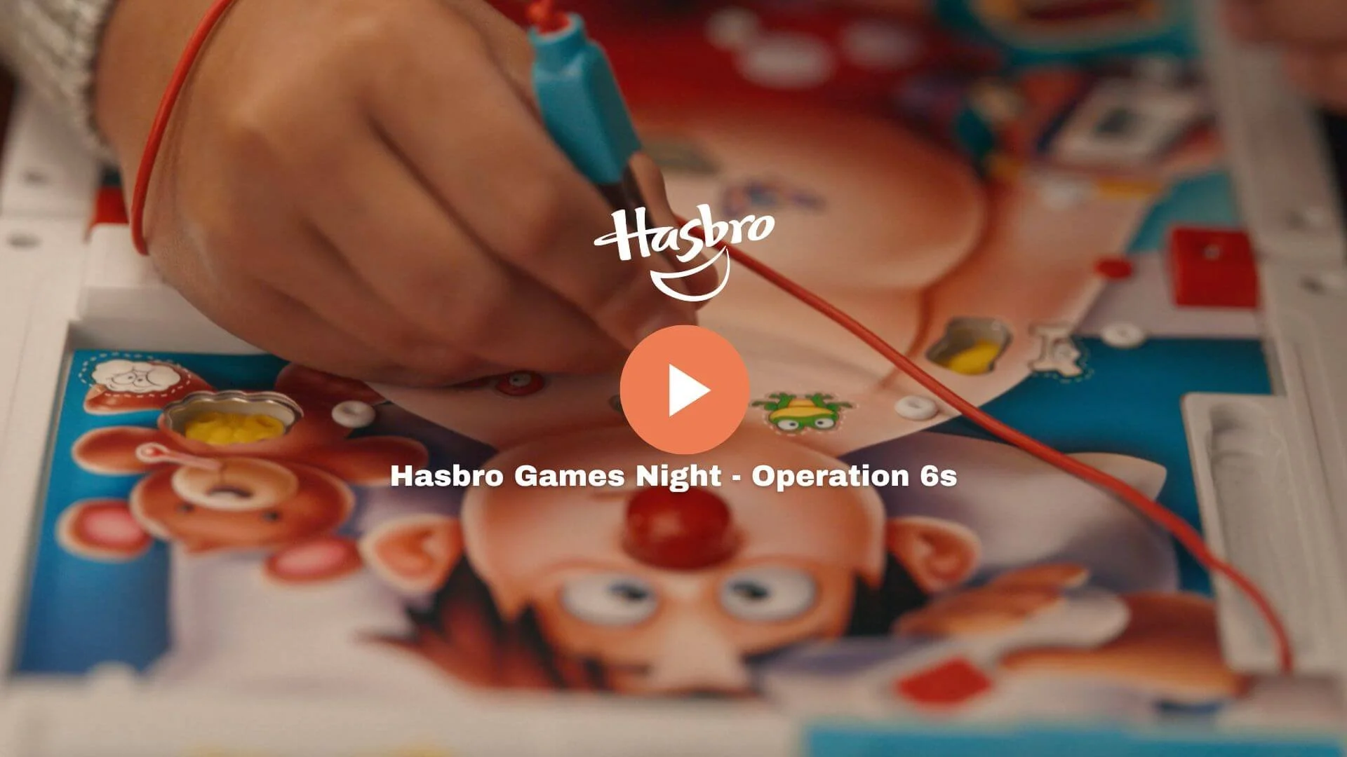 Close-up of someone holding a blue electrical test probe over a game board for Hasbro Games Night - Operation 6s, with visible colorful cartoon characters and game pieces.