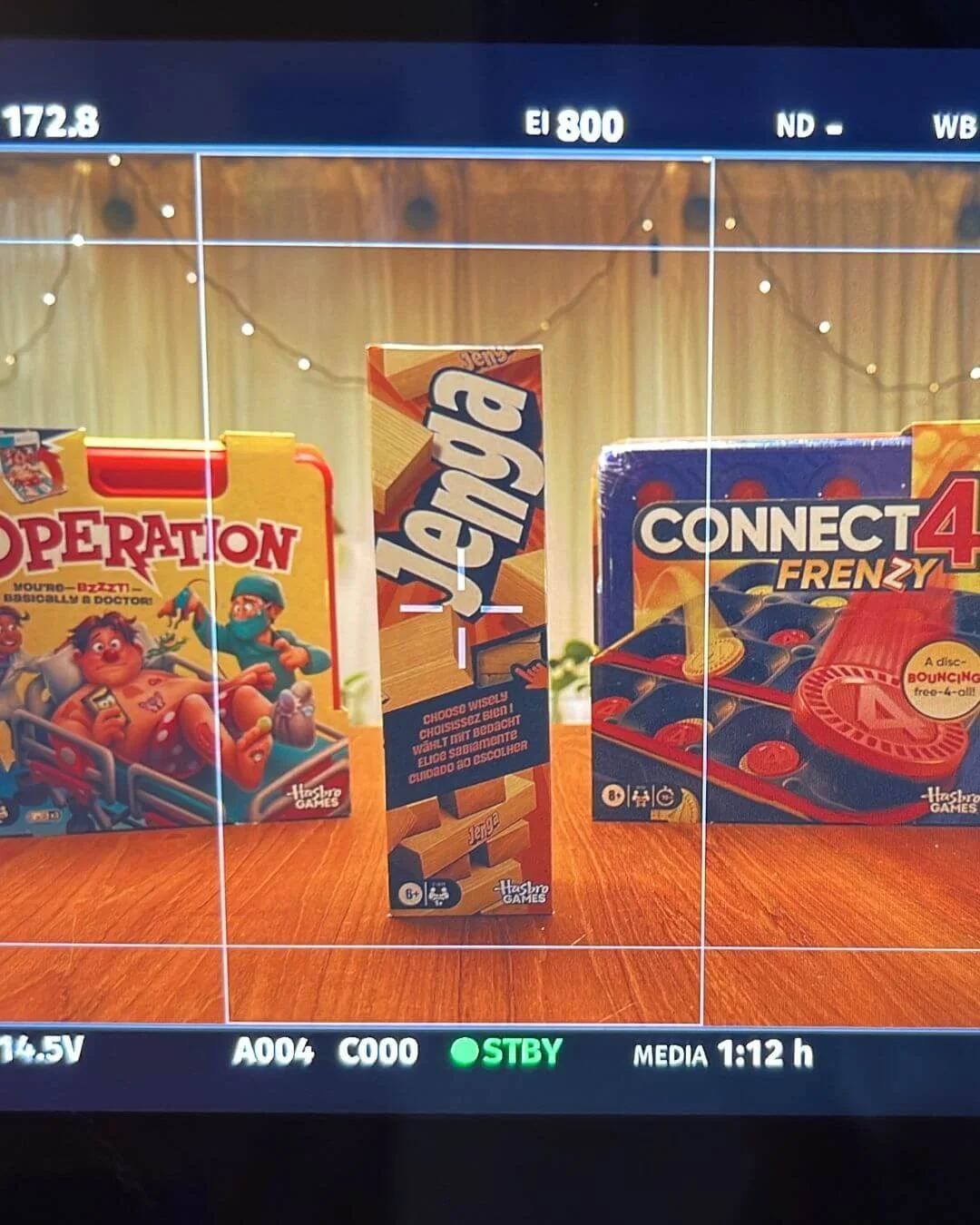 Photo of board games on a wooden table, including Operation, Jenga, and Connect 4 Frenzy, with a festive string light in the background.