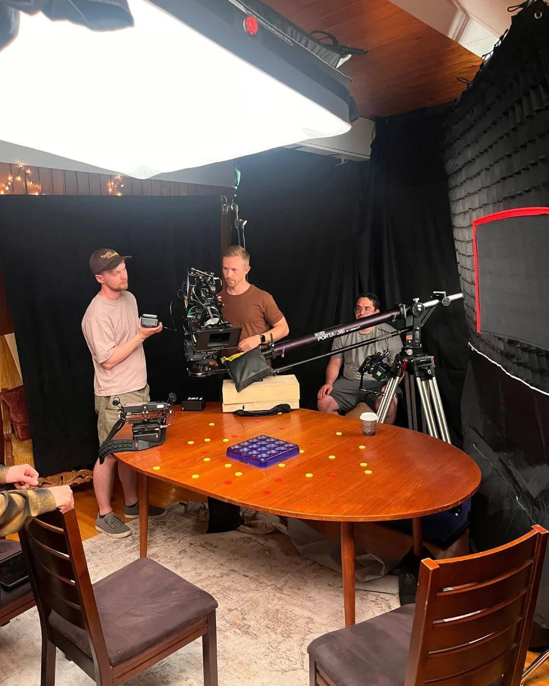 Filming crew setting up for a video shoot around a wooden dining table, with a camera mounted on a large rig, a person operating the camera, another person holding a box, and a third person sitting nearby in front of a black curtain backdrop in a dim