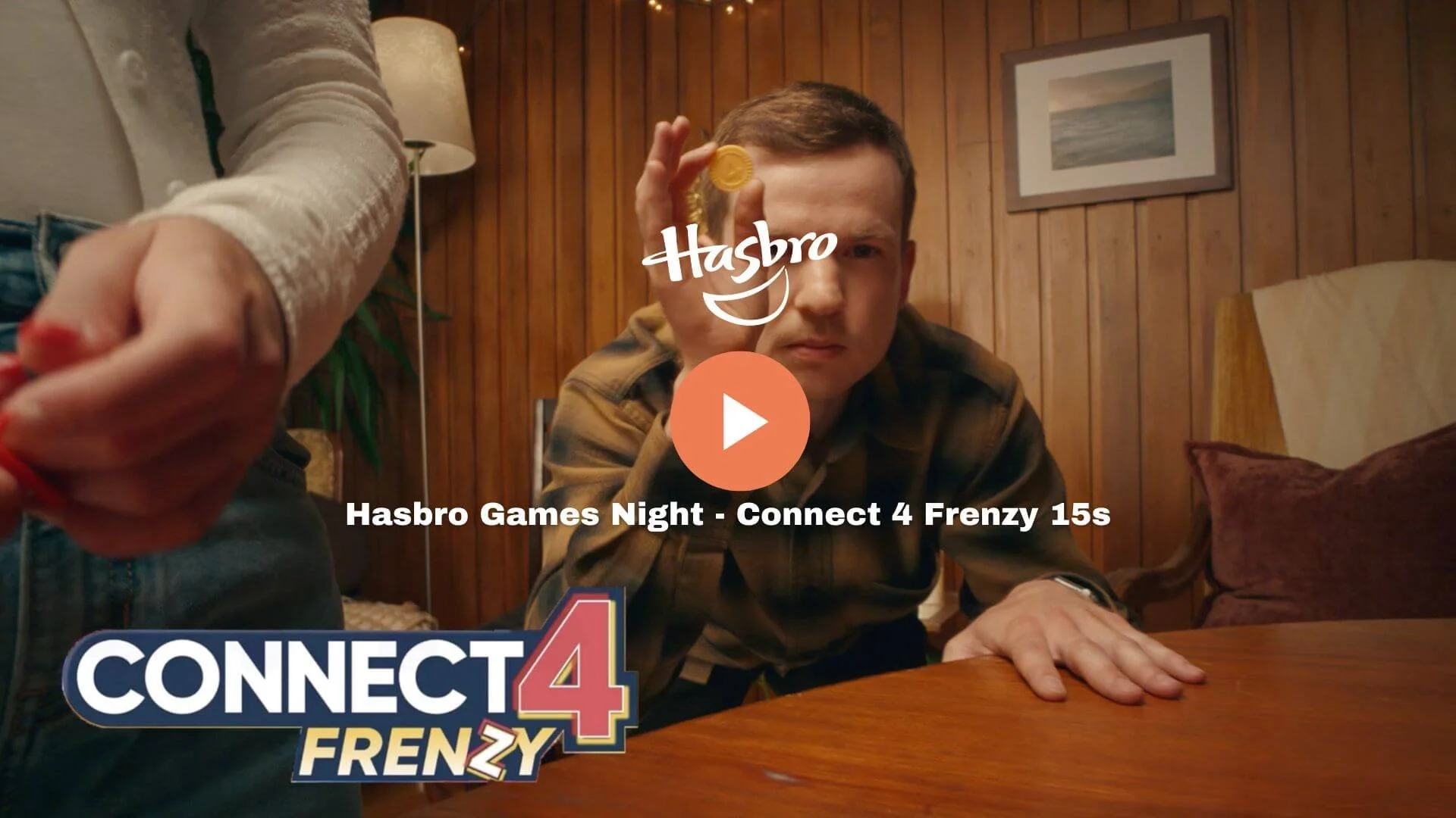 A young man in a plaid shirt, sitting at a wooden table with a wooden paneled wall behind him, holding a yellow game piece. A person's arm is reaching over the table, holding a red game piece. The image is overlaid with the words 'Hasbro' and 'Connect 4 Frenzy' logo, and text indicating it is from a video titled 'Hasbro Games Night - Connect 4 Frenzy 15s'.