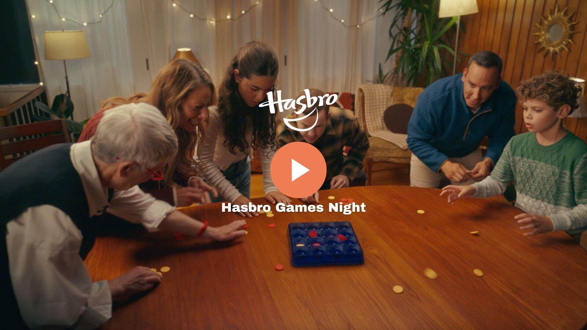 A group of people, including children and adults, gathered around a wooden table playing a game with colored discs, in a cozy room decorated with string lights, lamps, and a plant, during a Hasbro Games Night.