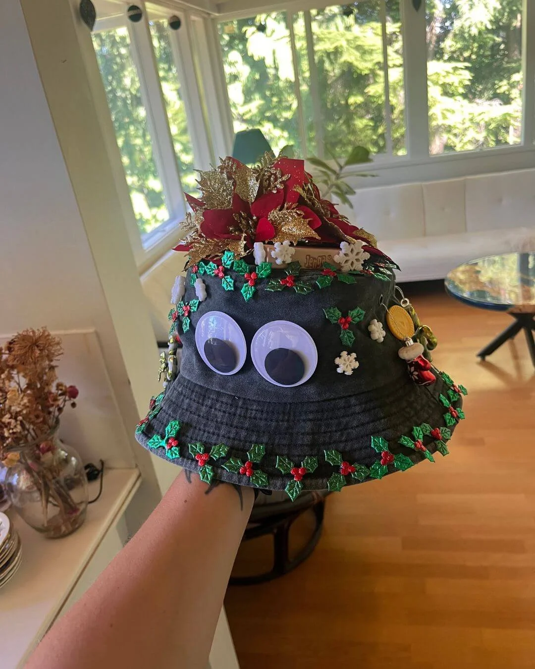 Festive Christmas hat decorated with googly eyes, holly leaves, red berries, gold and red flowers, snowflake ornaments, and other holiday decorations.