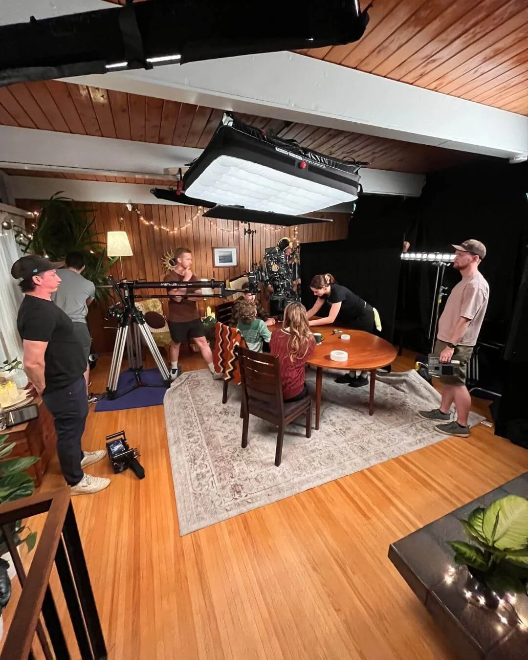 Film crew filming a scene around a dining table in a cozy room with wooden paneling, with equipment and crew members operating cameras and lights.