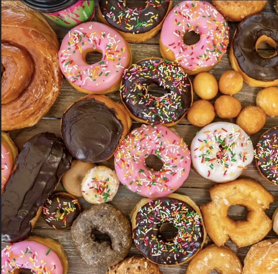An assortment of donuts decorated with colorful sprinkles, glazed icing in pink and white, and chocolate, arranged on a wooden surface along with a few small round pastries.