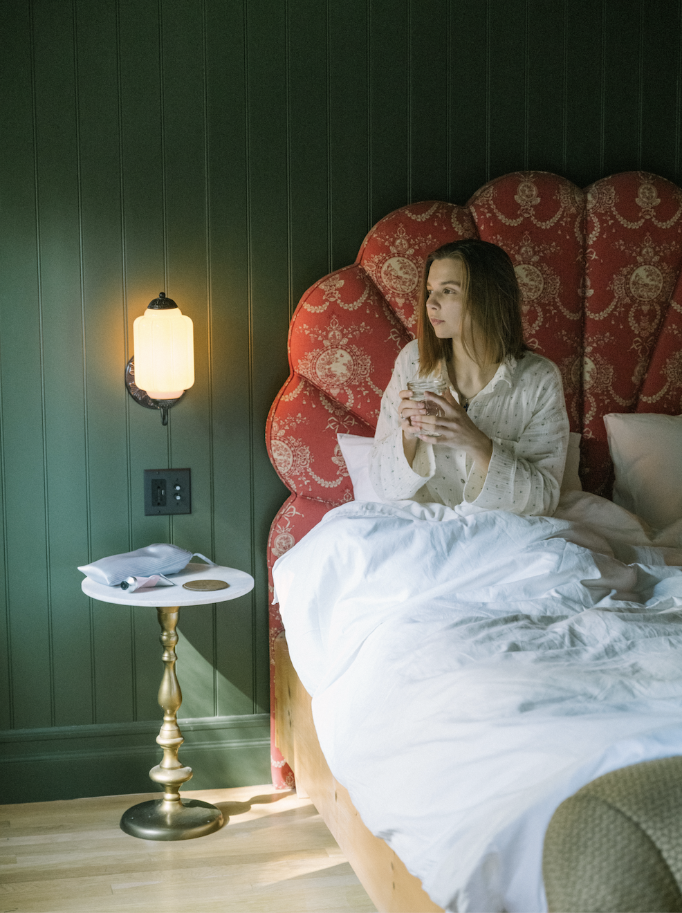 A woman sitting in bed holding a glass, with a green wall, a side table with a spiral base, a lamp, a wall outlet, and some items on the table.
