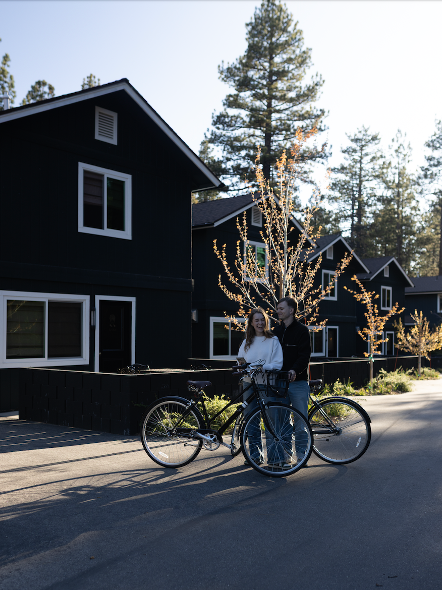 A couple standing with bikes in front of black residential buildings with lit trees in the background during daytime.