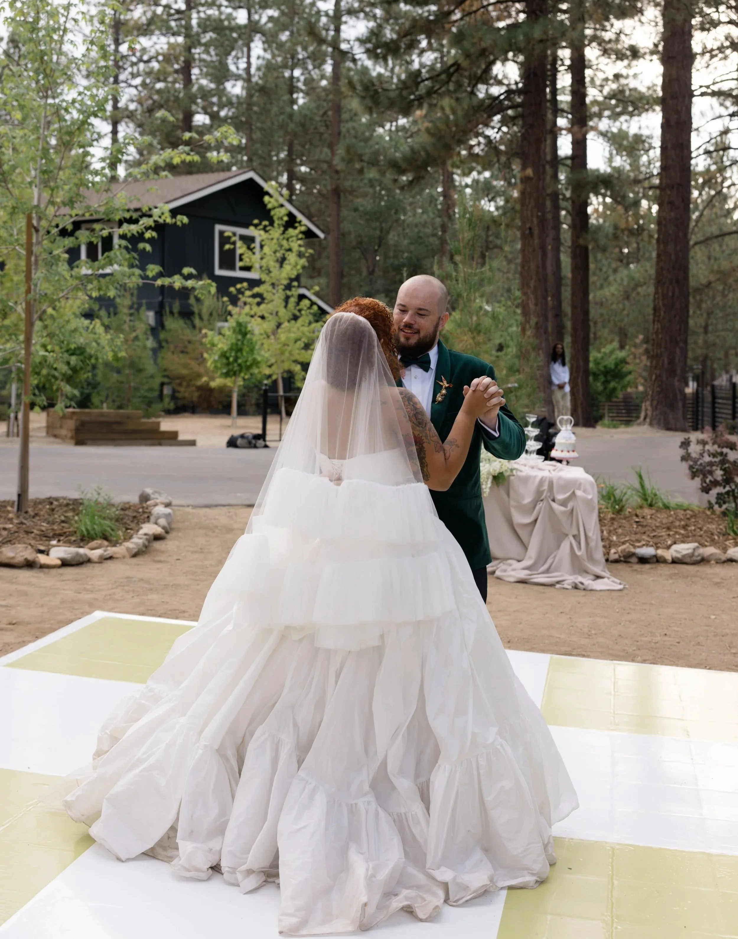 A couple dancing outdoors at a wedding ceremony, surrounded by trees with a black house in the background.