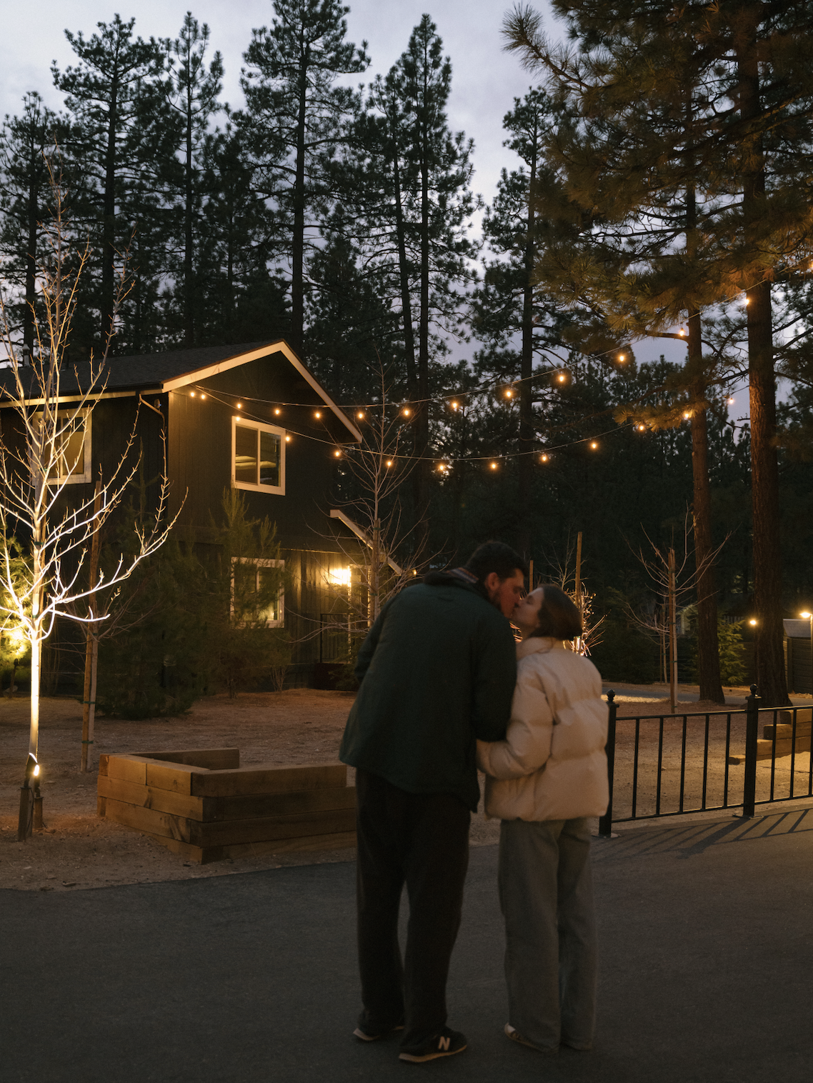 A couple sharing a kiss outdoors during dusk, with string lights overhead, trees, and a black house with lit windows in the background.