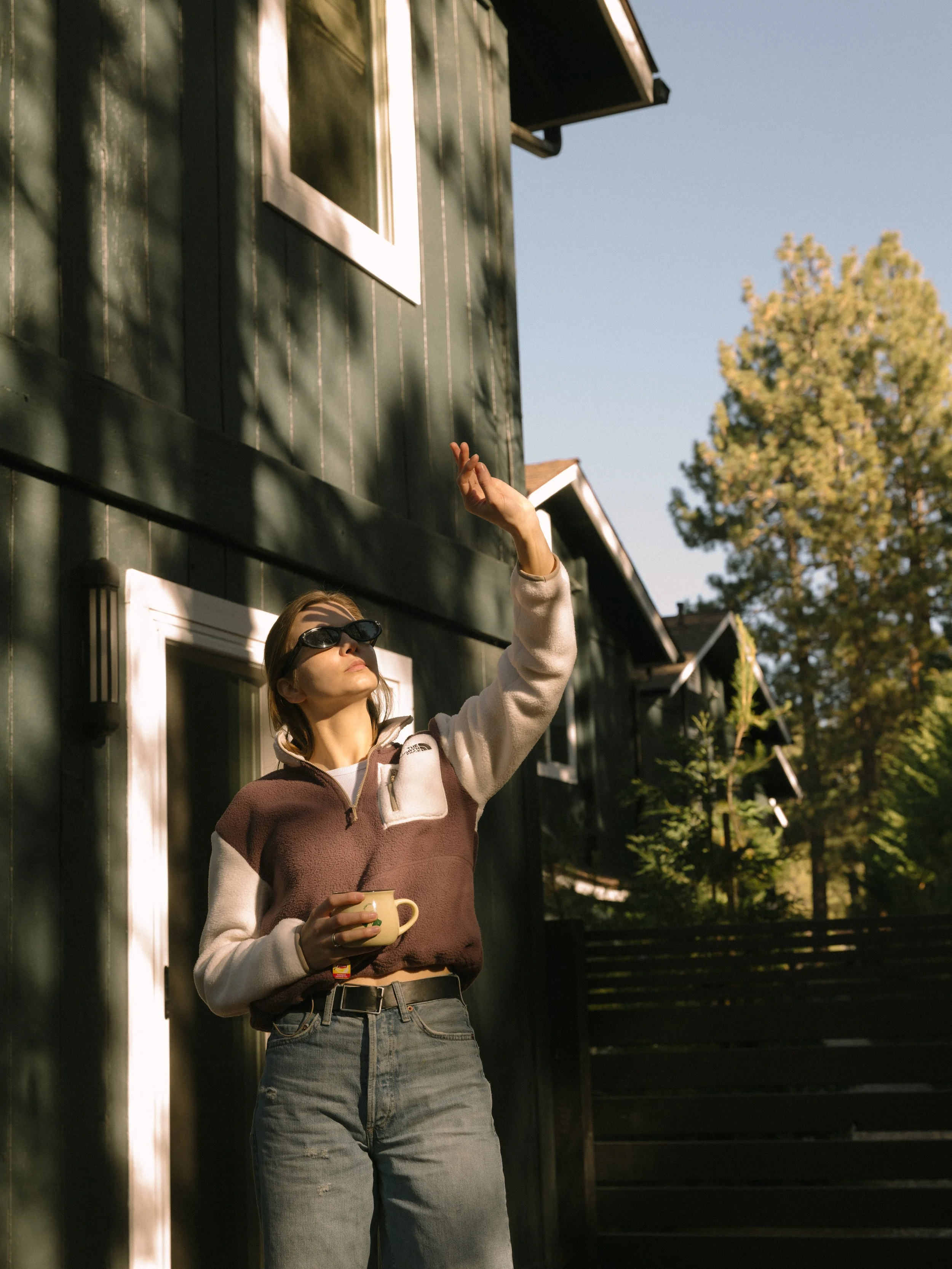 A woman with sunglasses and casual clothes is standing outside a green house, raising her hand and holding a yellow mug, with her face turned upward.