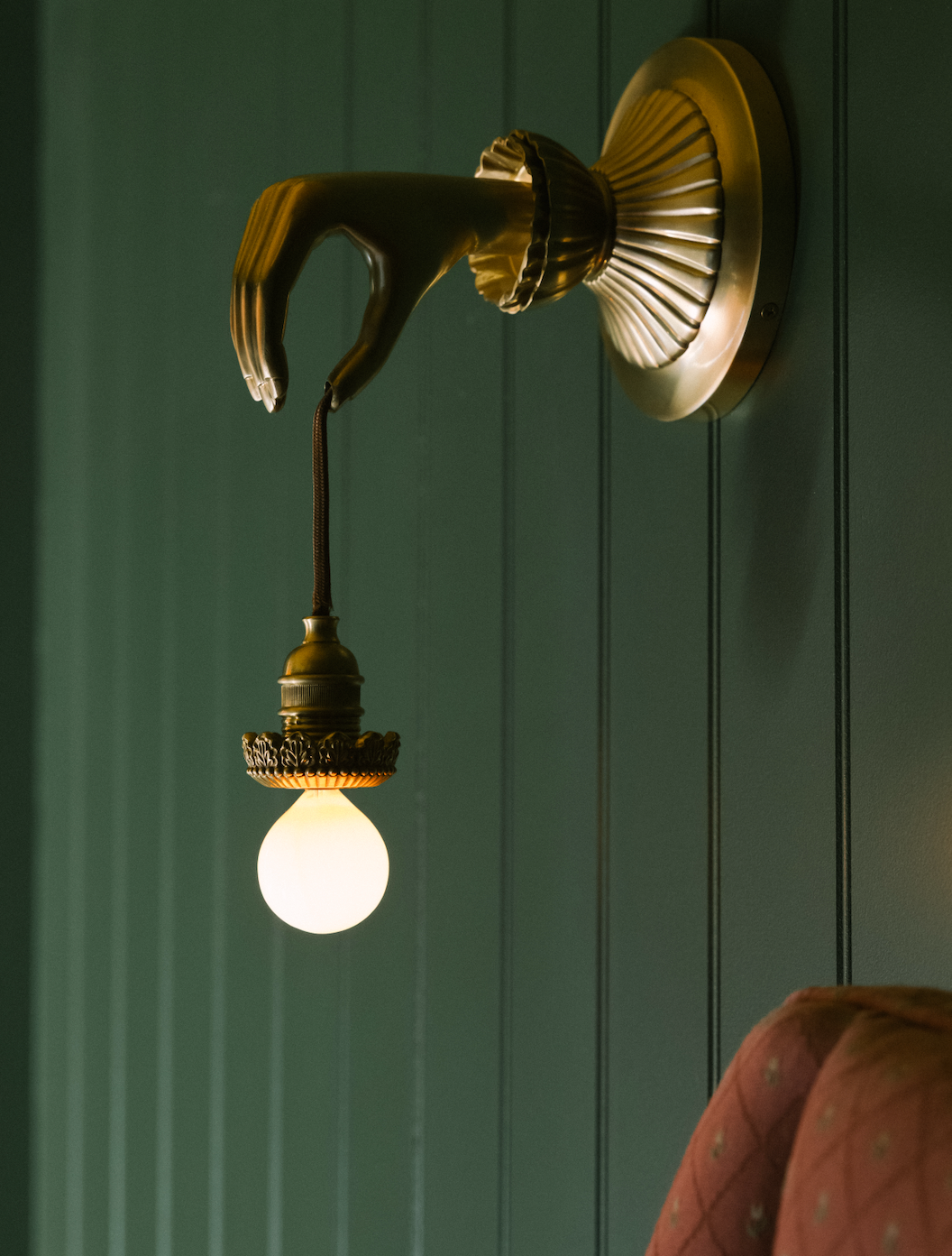 Decorative wall-mounted light fixture with a hand-shaped design, featuring a light bulb hanging from the fingers, mounted on a green paneled wall.