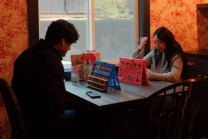 A man and a woman sitting at a table in a restaurant or cafe, playing a game with red and blue boards that have numbers and symbols, with items like drinks and a phone on the table, near a window with curtains.