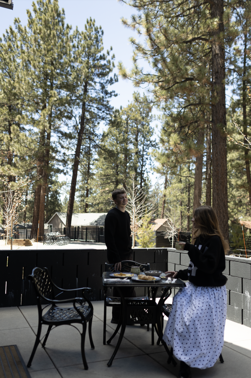 People having breakfast outdoors on a patio surrounded by tall pine trees and blue sky.
