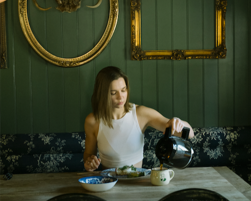 A young woman in a white sleeveless top is pouring coffee from a black coffee pot into a mug at a dining table. The table has plates and a teapot on it. The background features dark green paneling with empty gold picture frames hanging on the wall.