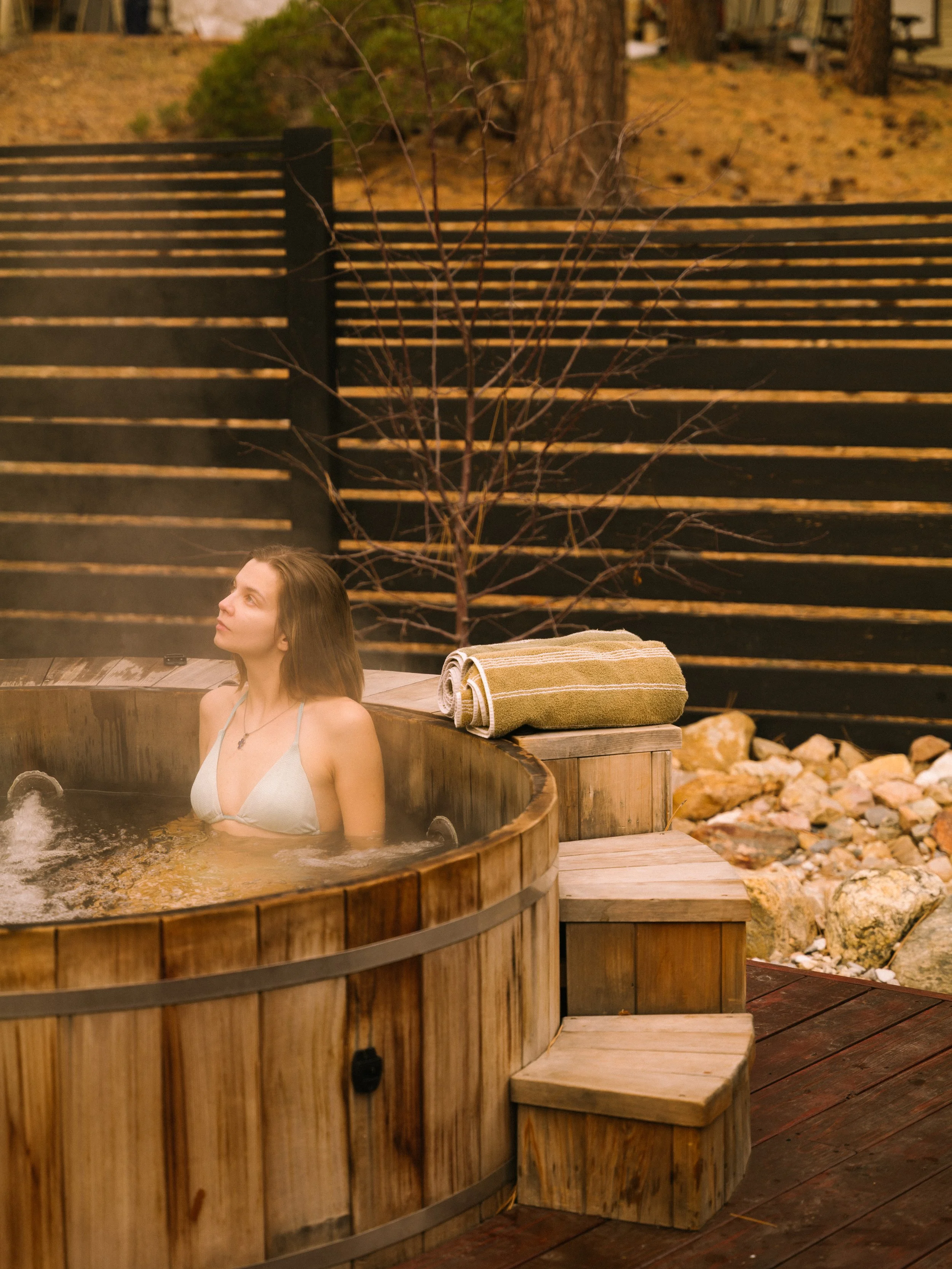 A woman with long brown hair is sitting in a wooden hot tub filled with water, outdoors during a cold season. She is gazing upward and has a towel and a small object on the edge of the hot tub. The background features a black wooden fence, a leafless tree, and a forest with pine trees and rocks.