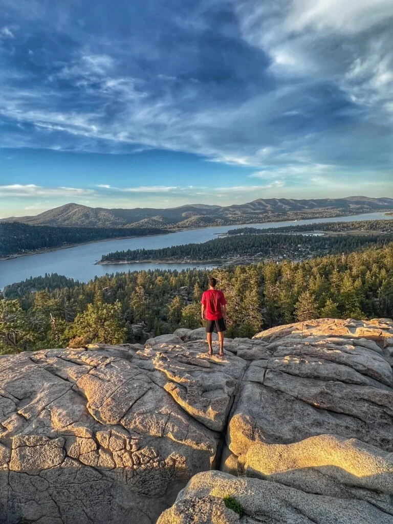A person in a red shirt and shorts standing on a rocky outcrop overlooking a landscape with a lake, forest, and mountains under a partly cloudy sky.