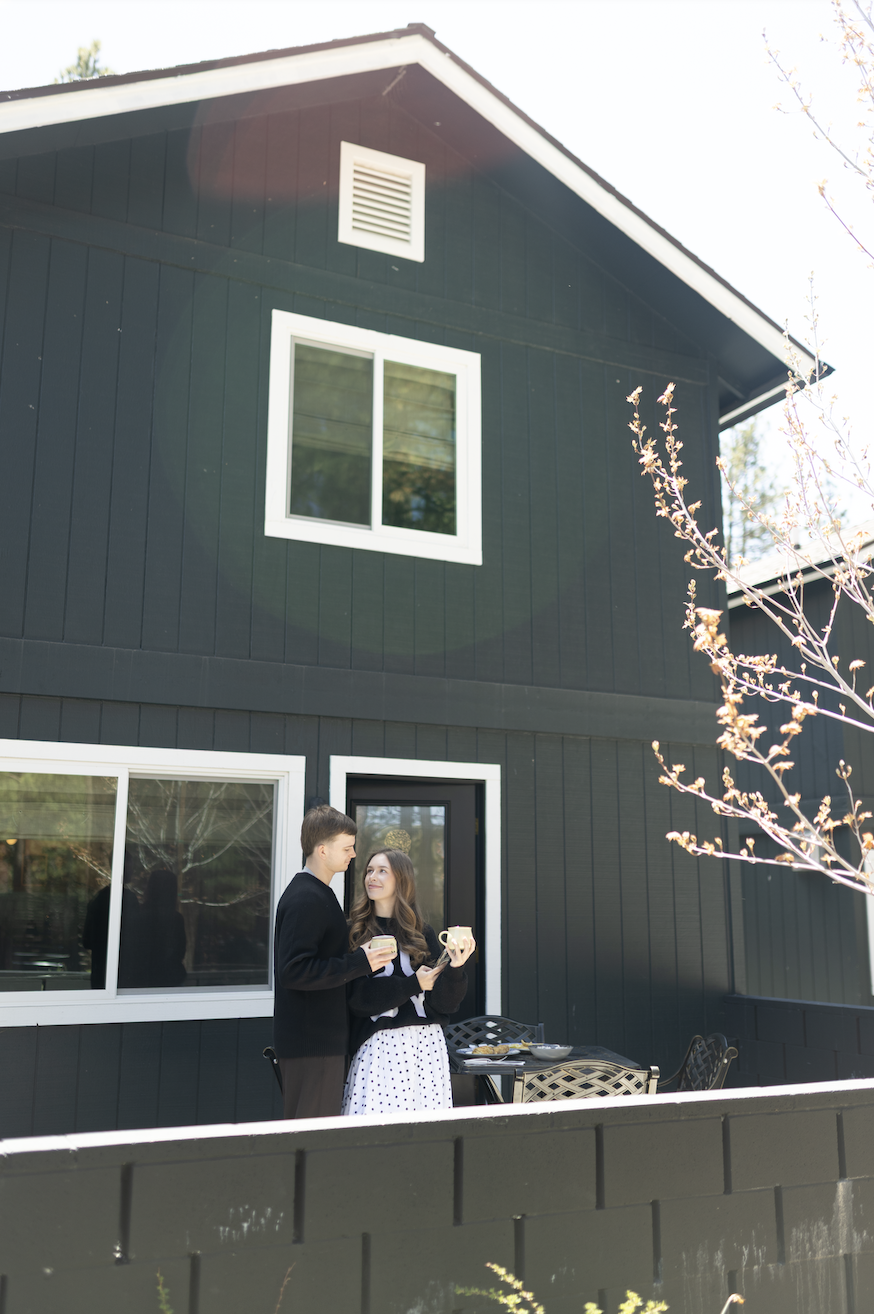 Two people standing on a porch in front of a dark-colored house, holding cups and enjoying a conversation. The house has white-framed windows and a door, with some outdoor furniture on the porch.