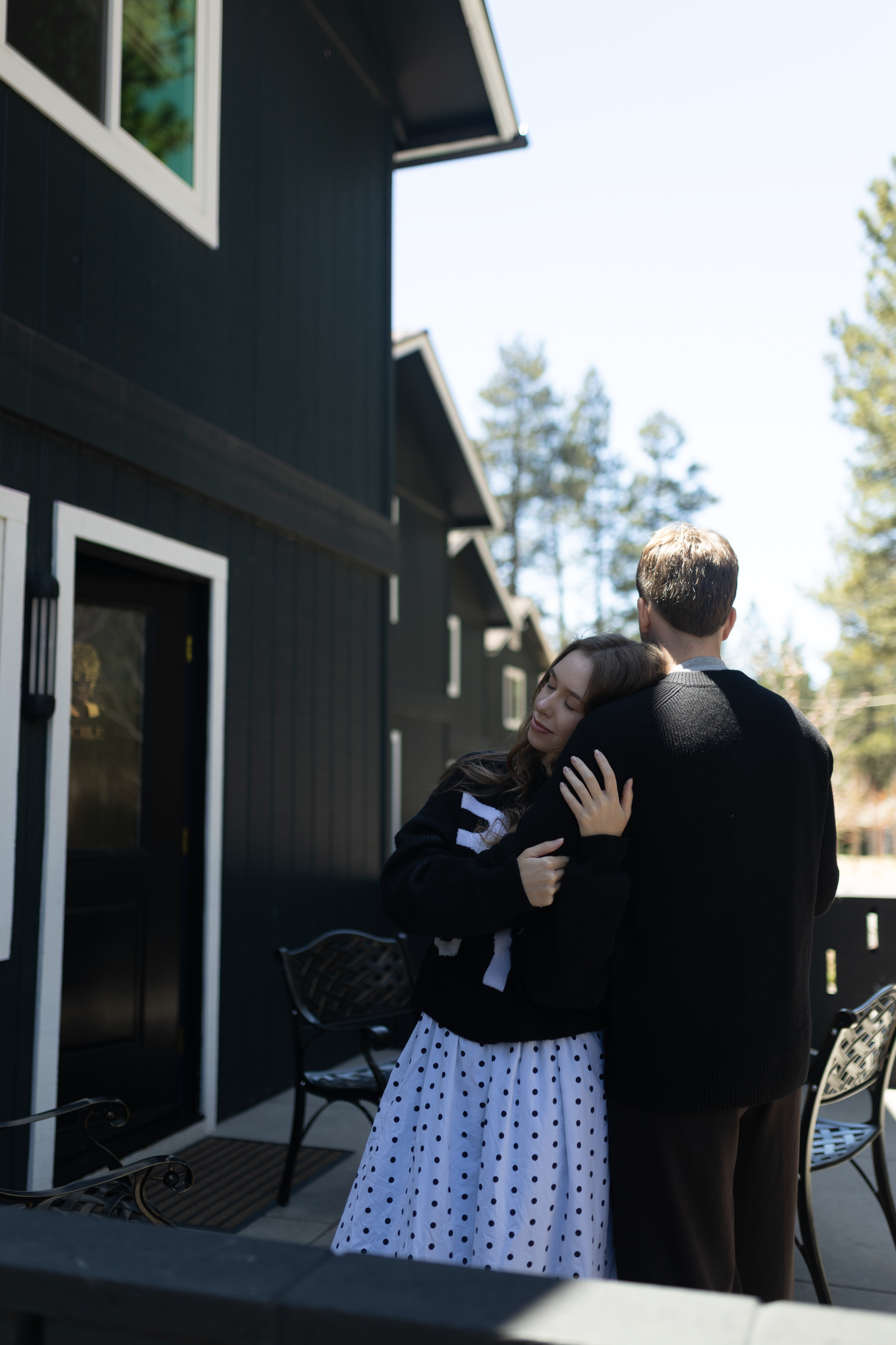 A woman with long dark hair and a man with short light brown hair hugging outside a dark-colored house with white trim on a sunny day.