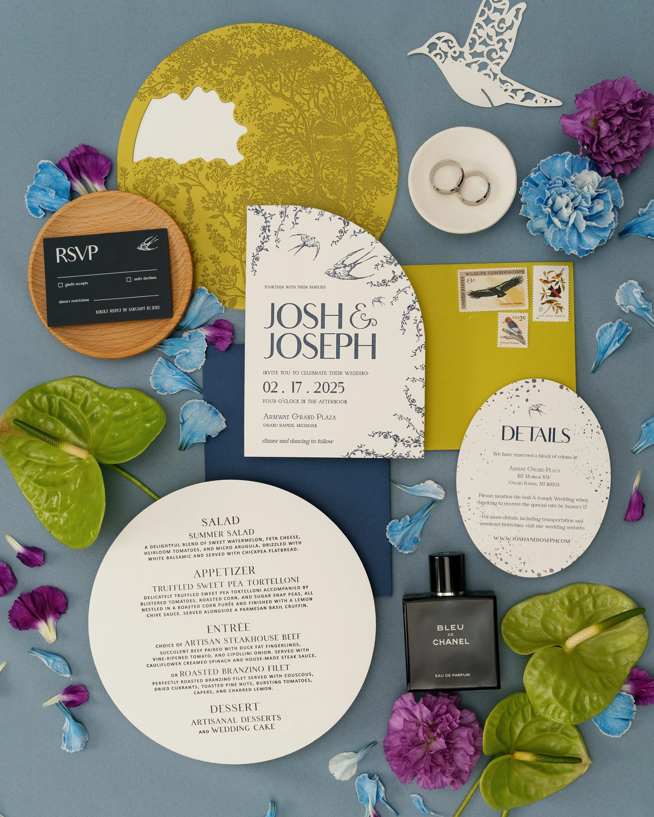 Wedding invitation suite with details, RSVP, and menu cards, surrounded by flowers, a caller, stamps, a perfume bottle, wedding bands, and decorative paper cutouts of hummingbirds and a bird in flight on a blue background.