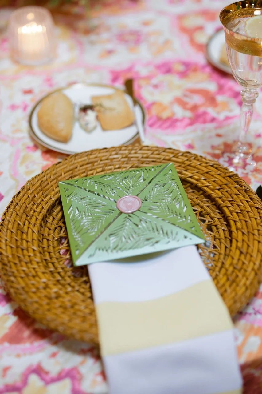 Table setting with a woven placemat, a white and yellow napkin, a green palm leaf cutout greeting card, a partially eaten dessert on a plate, a glass of white wine, and a candle in the background with floral tablecloth.