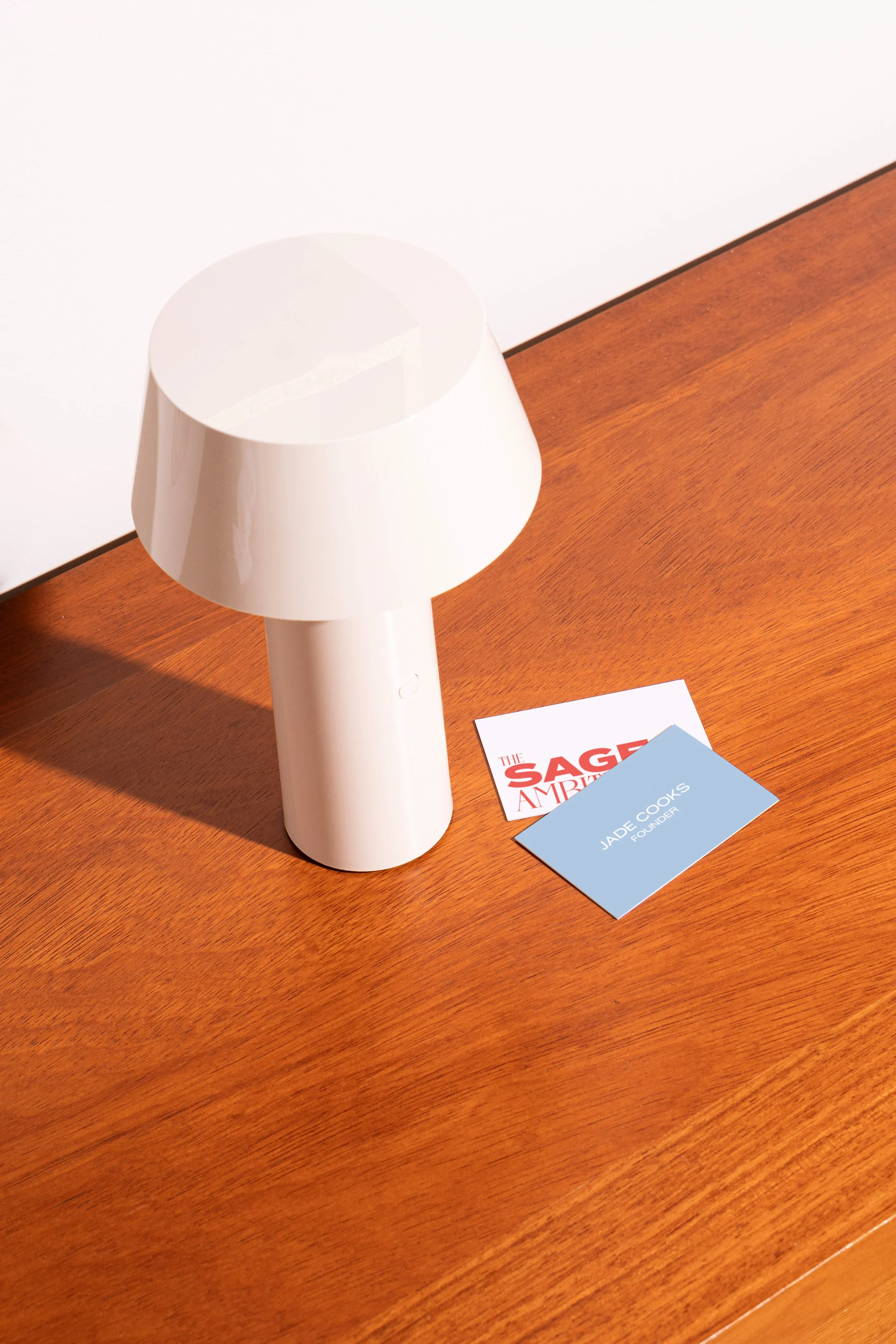 A white lamp with a rounded shade on a wooden surface next to two business cards, one red and white and the other blue, with the name Jade Cooks and the title Founder.