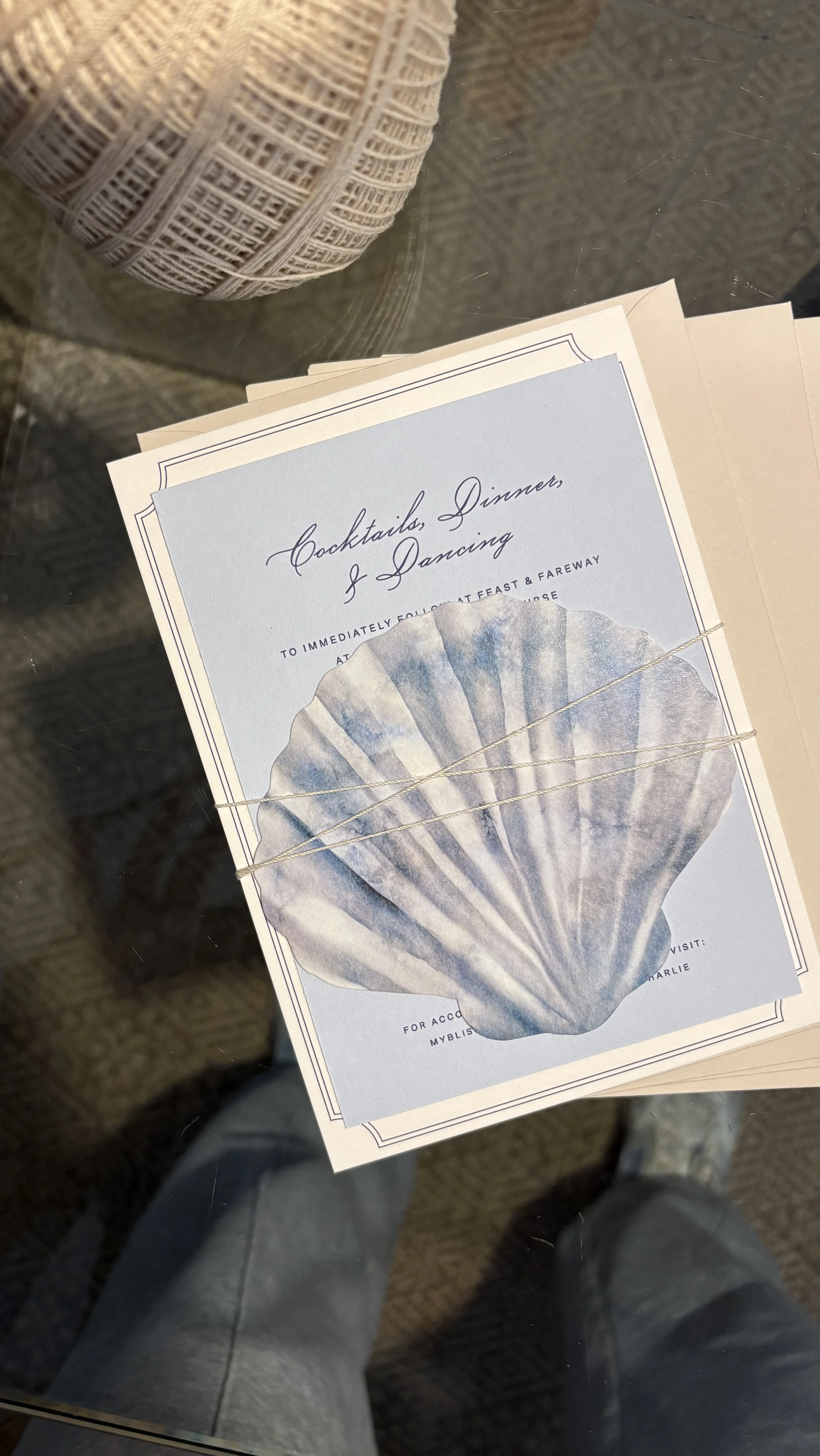 A stack of event invitations with a seashell design, tied with a piece of string, resting on a reflective glass surface. A large ball of neutral-colored yarn is visible in the background.