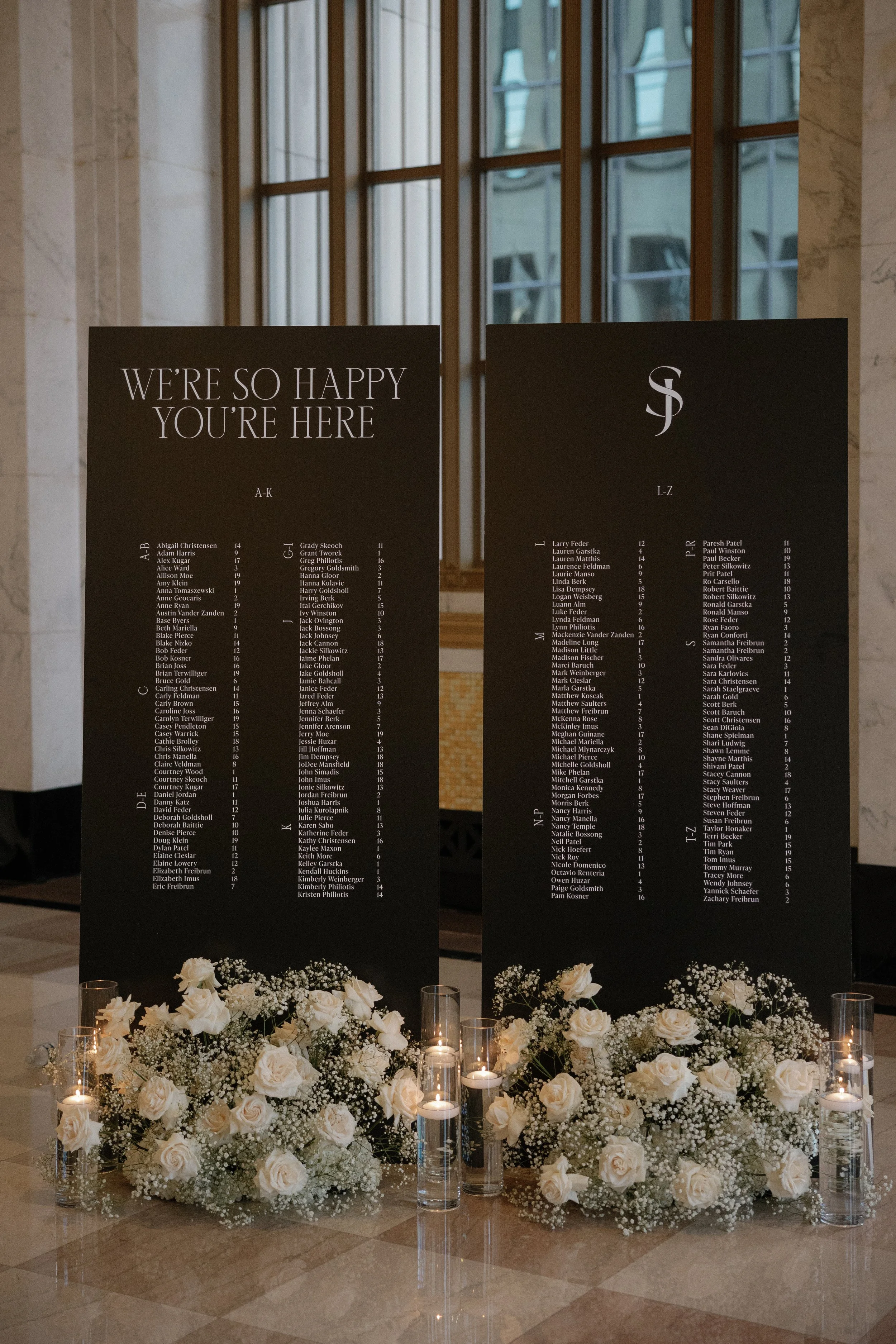 Wedding seating chart displays two large black boards with guest names grouped alphabetically, decorated with white roses, baby's breath, and candles at the base.