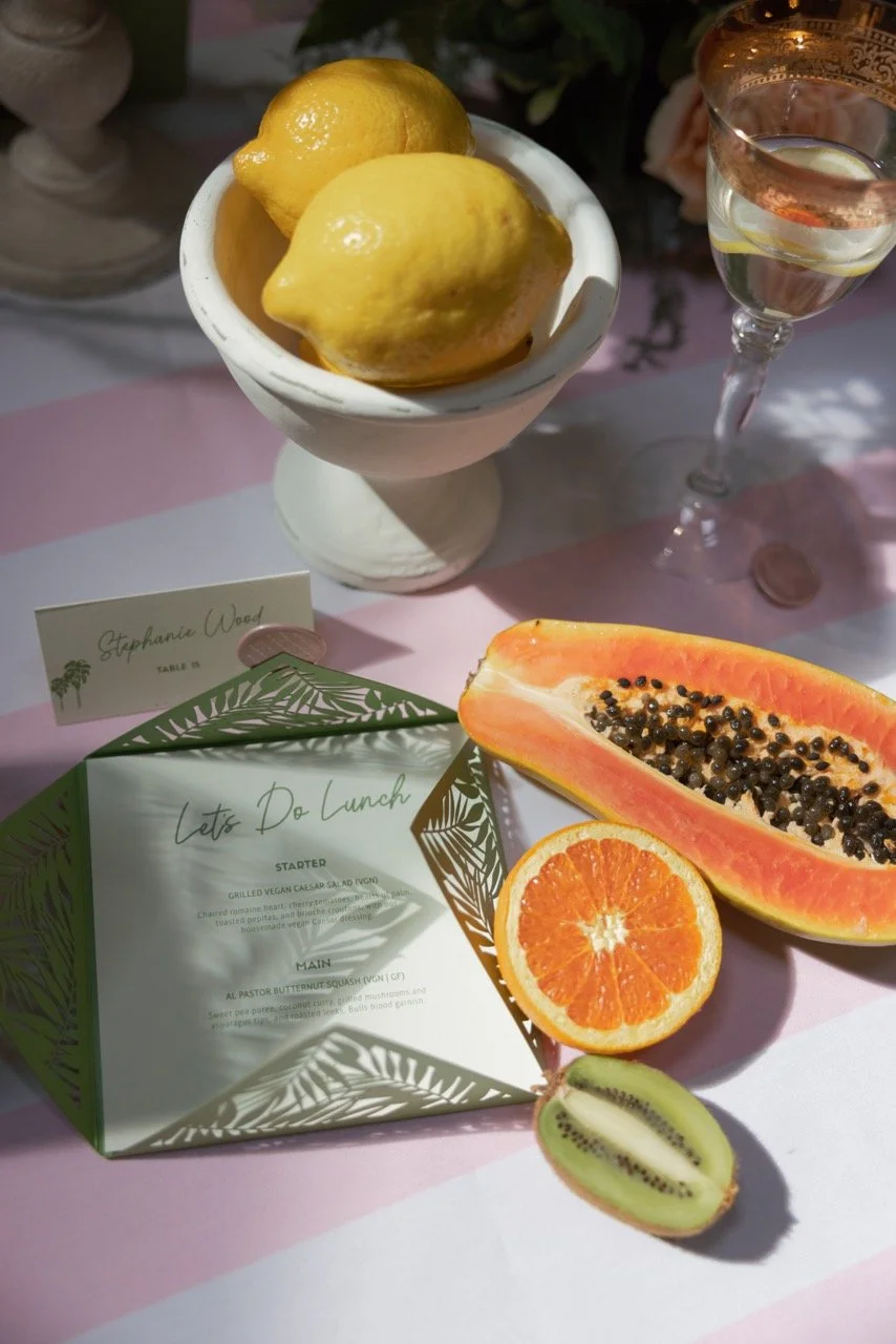 Lemon in a white cup, sliced orange, halved kiwi, halved papaya with black seeds, a glass of water with lemon, and a place card reading 'Stephanie Wood' on a table.